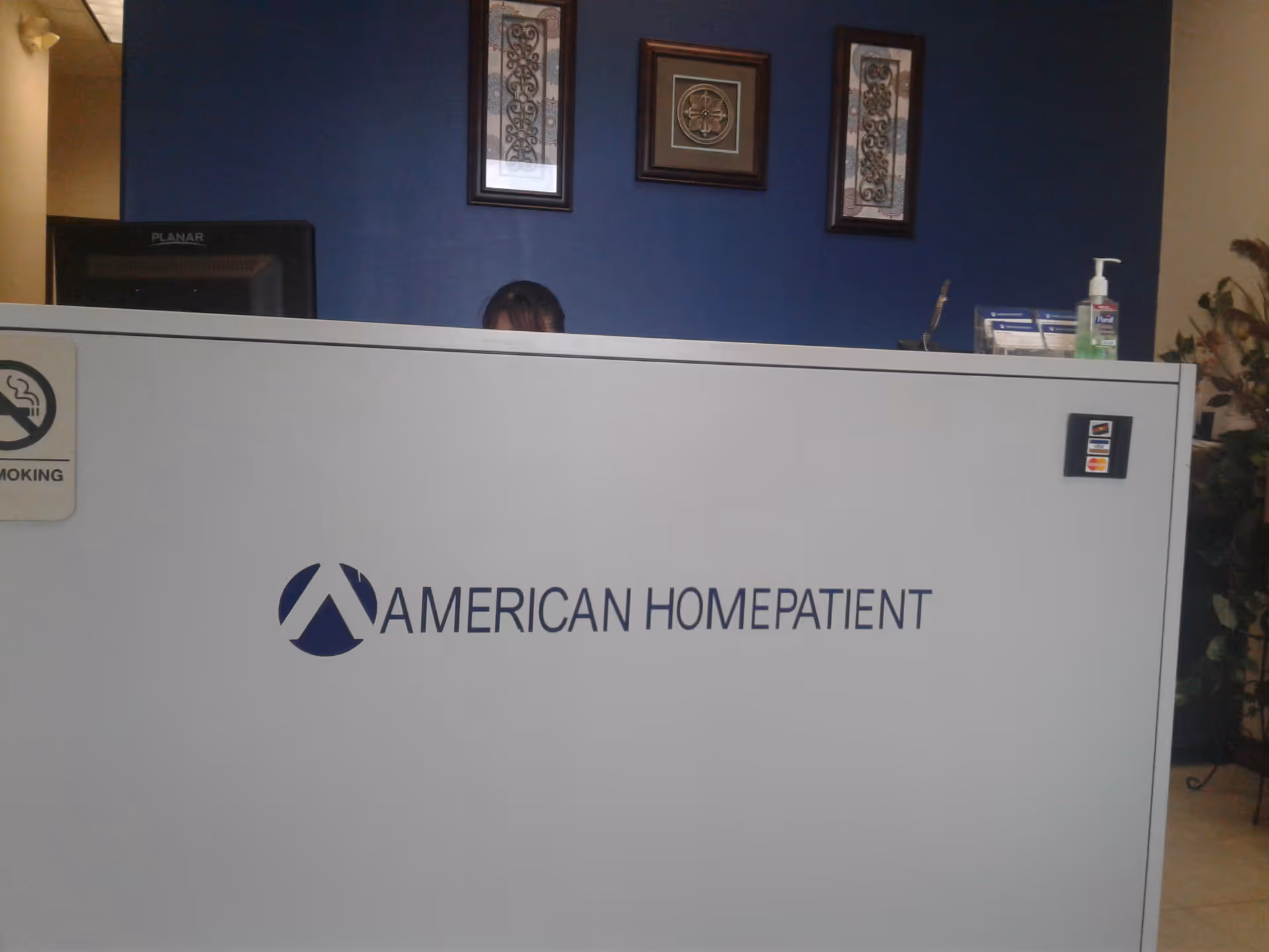Reception desk with American HomePatient logo and blue wall, partial person behind desk.