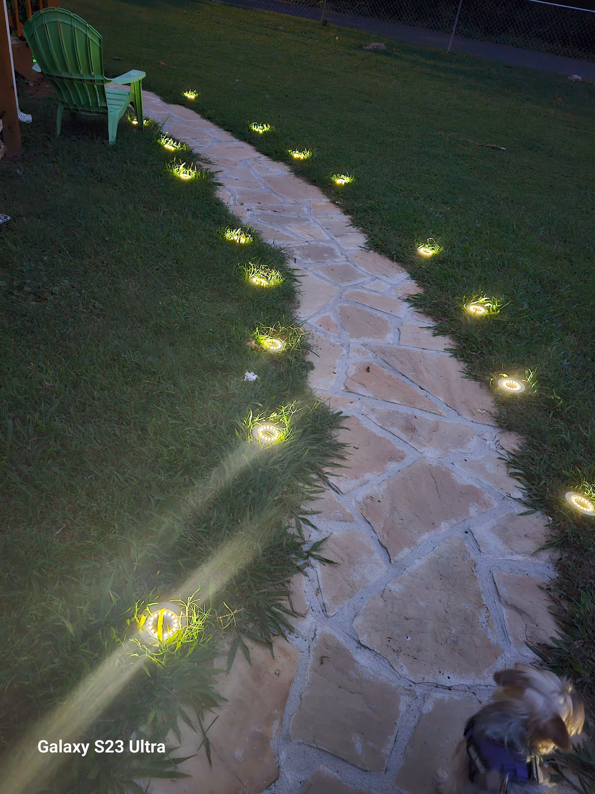 Stone path through a grassy yard with illuminated ground lights and a green chair.