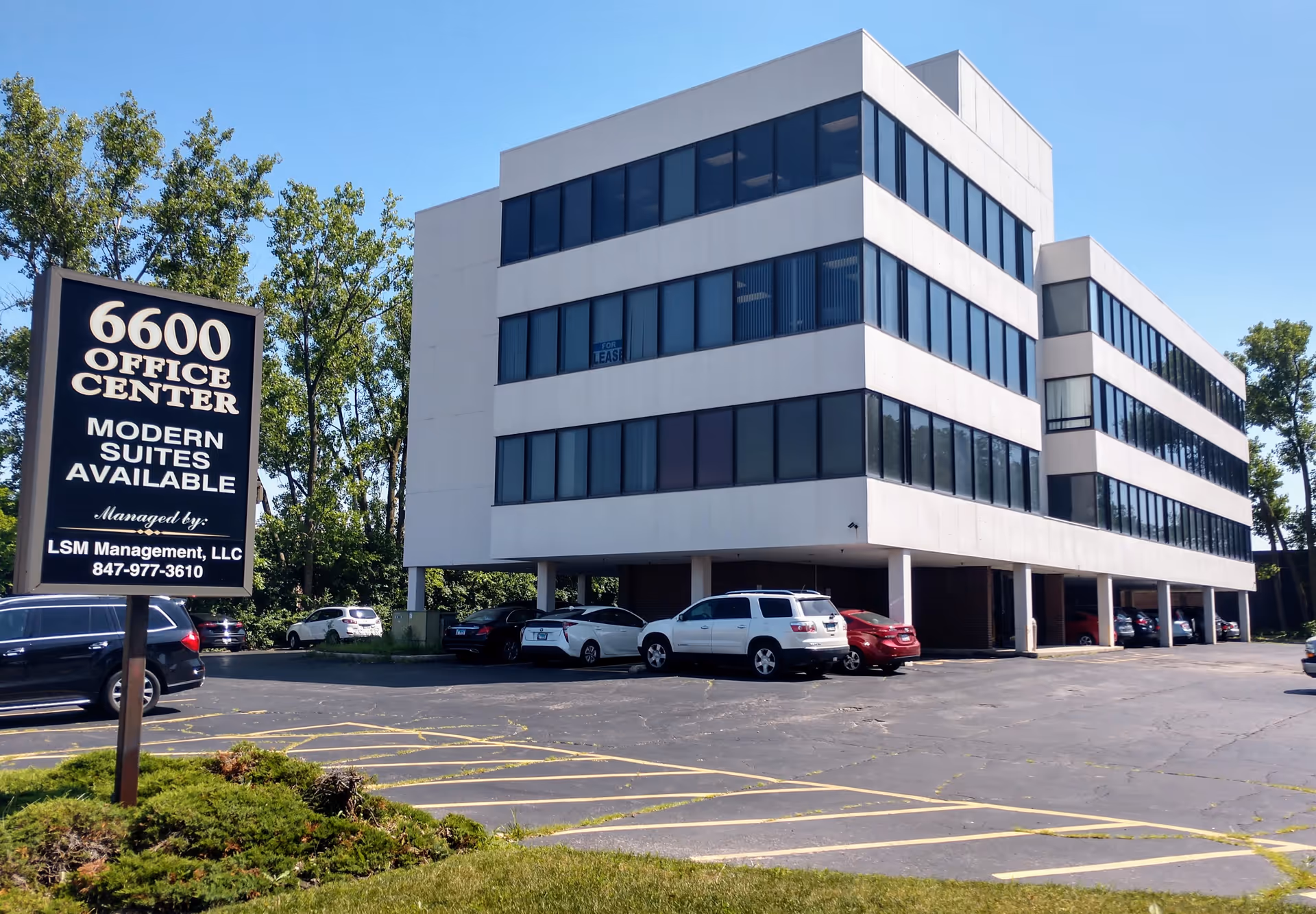 A white, modern, multi-story office building with a parking lot and signboard in front.