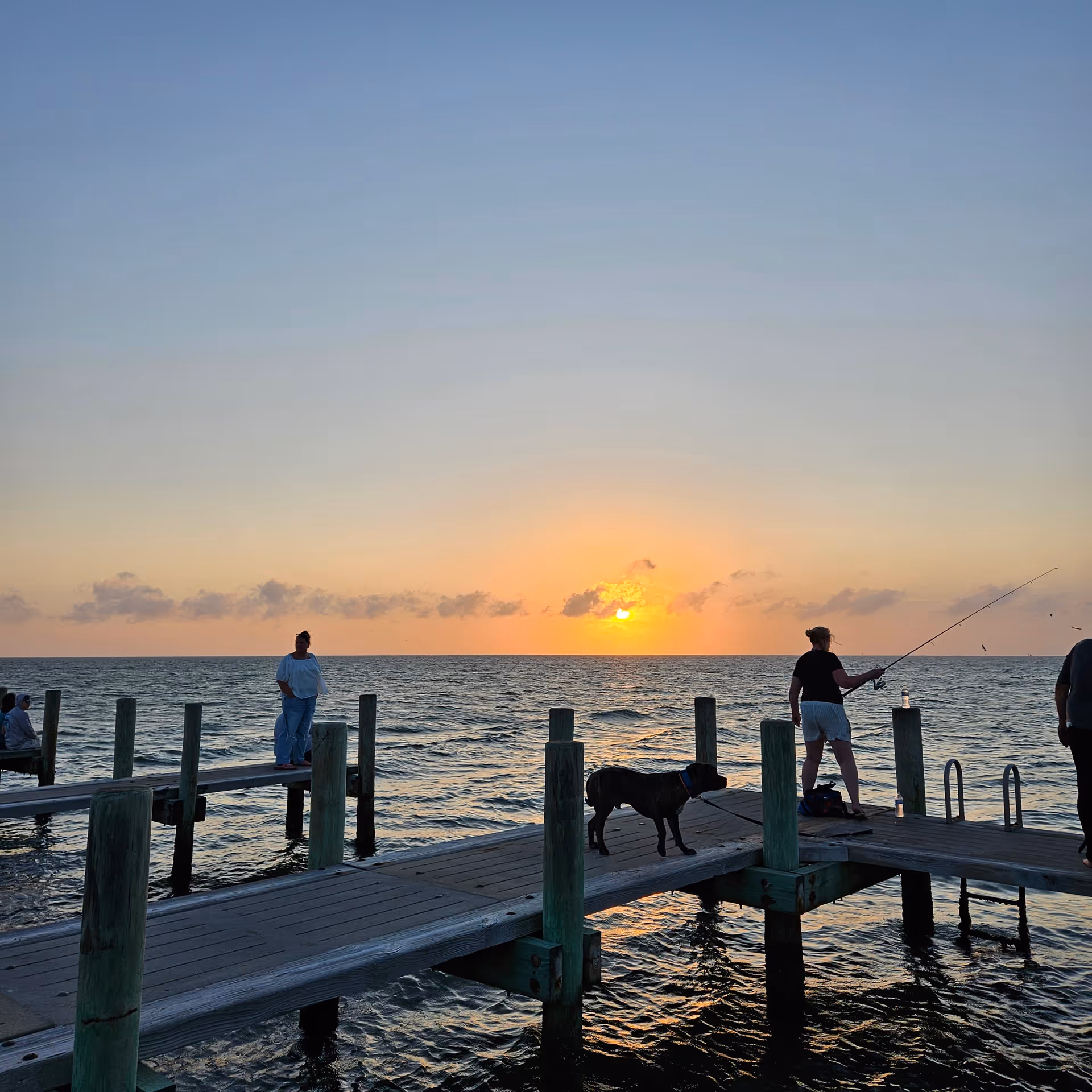 Sunset over a wooden pier with a dog and people fishing by the calm sea.