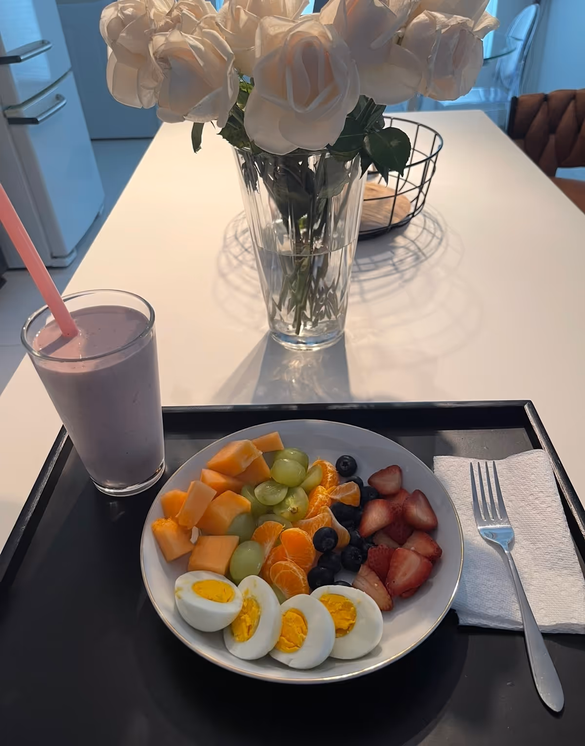 A white table features a fruit plate, smoothie, and a vase with white roses.