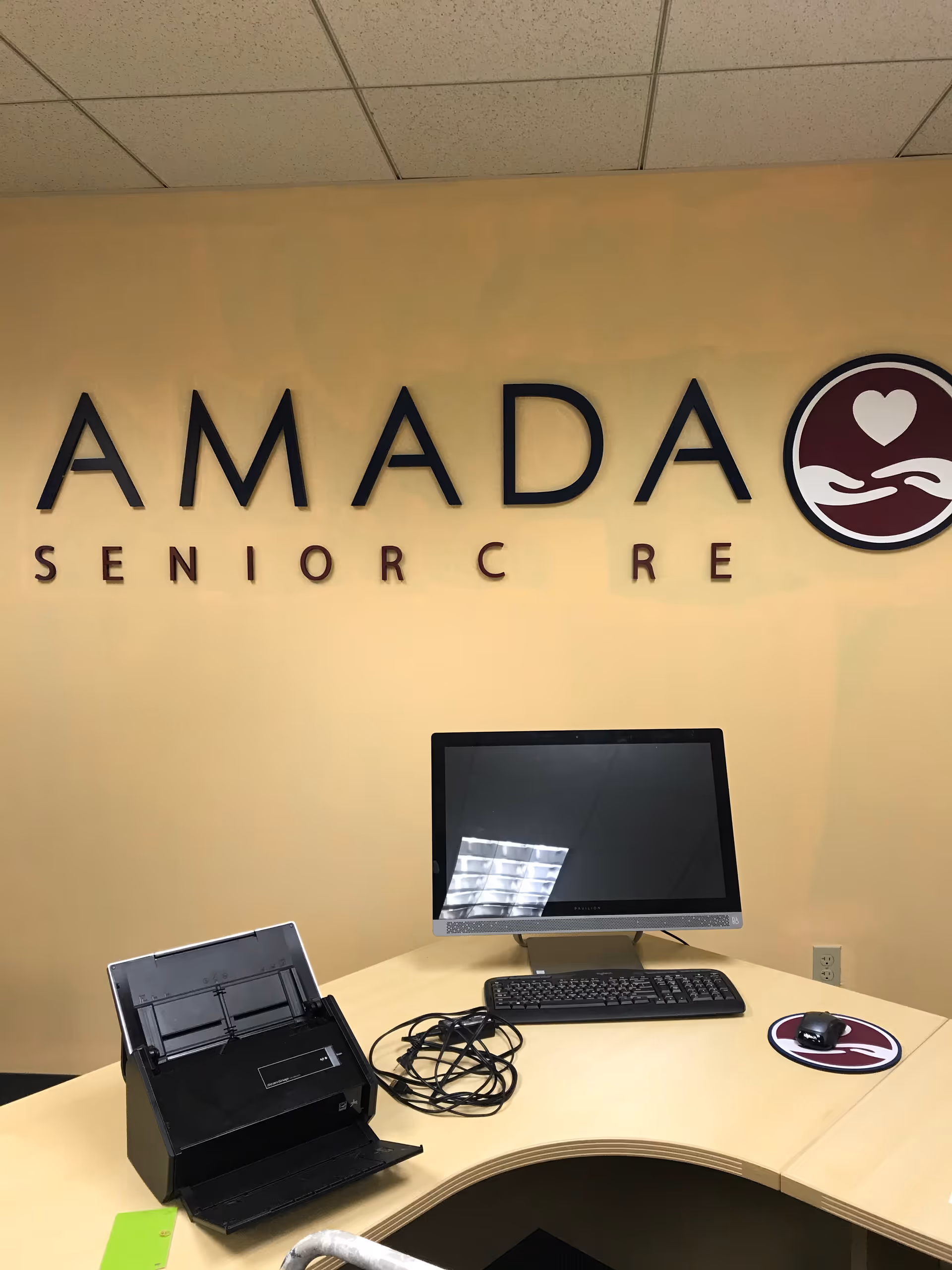 Reception desk with computer, printer, and Amada Senior Care logo on wall.