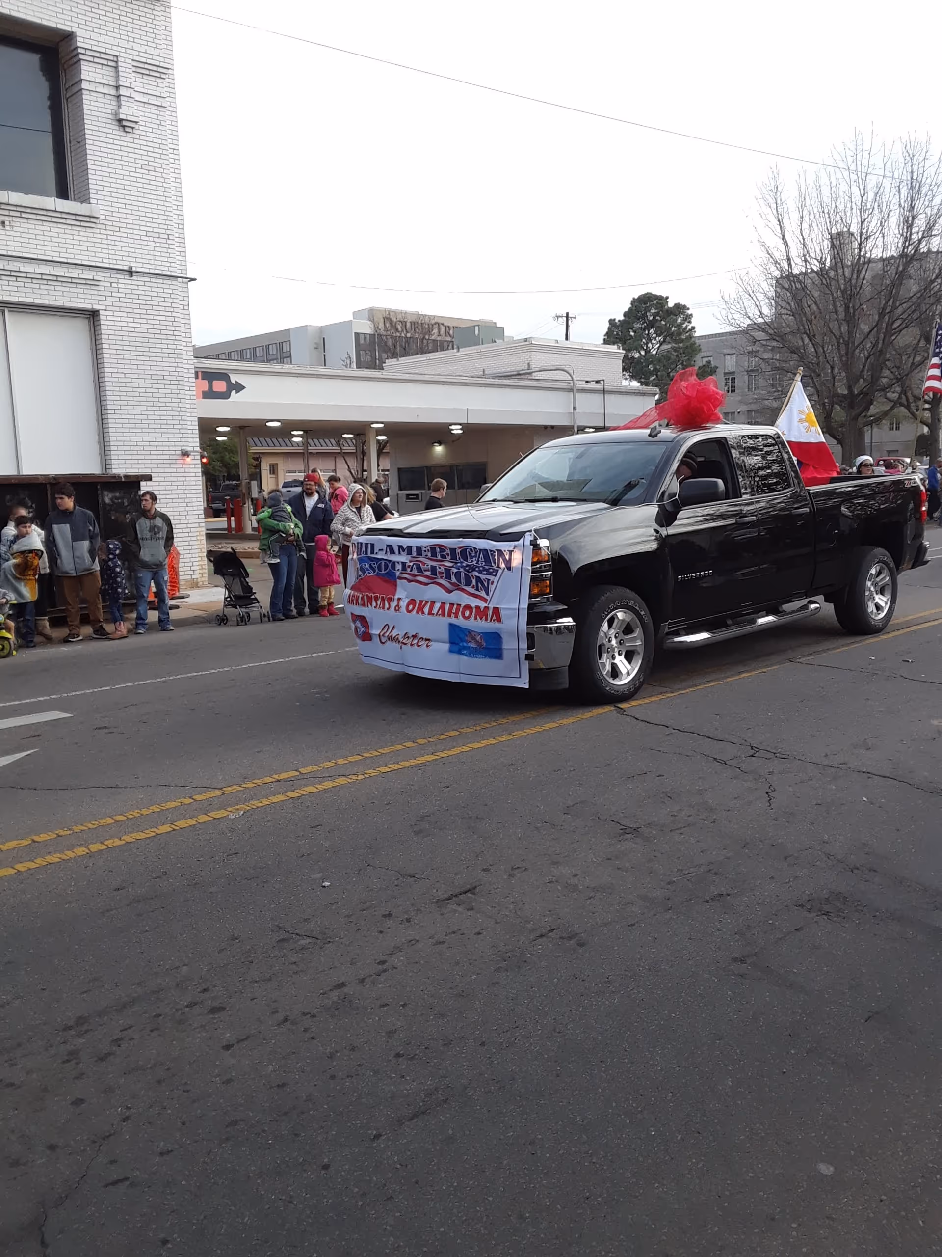 Black pickup truck with a banner drives down a parade street; crowd watches.