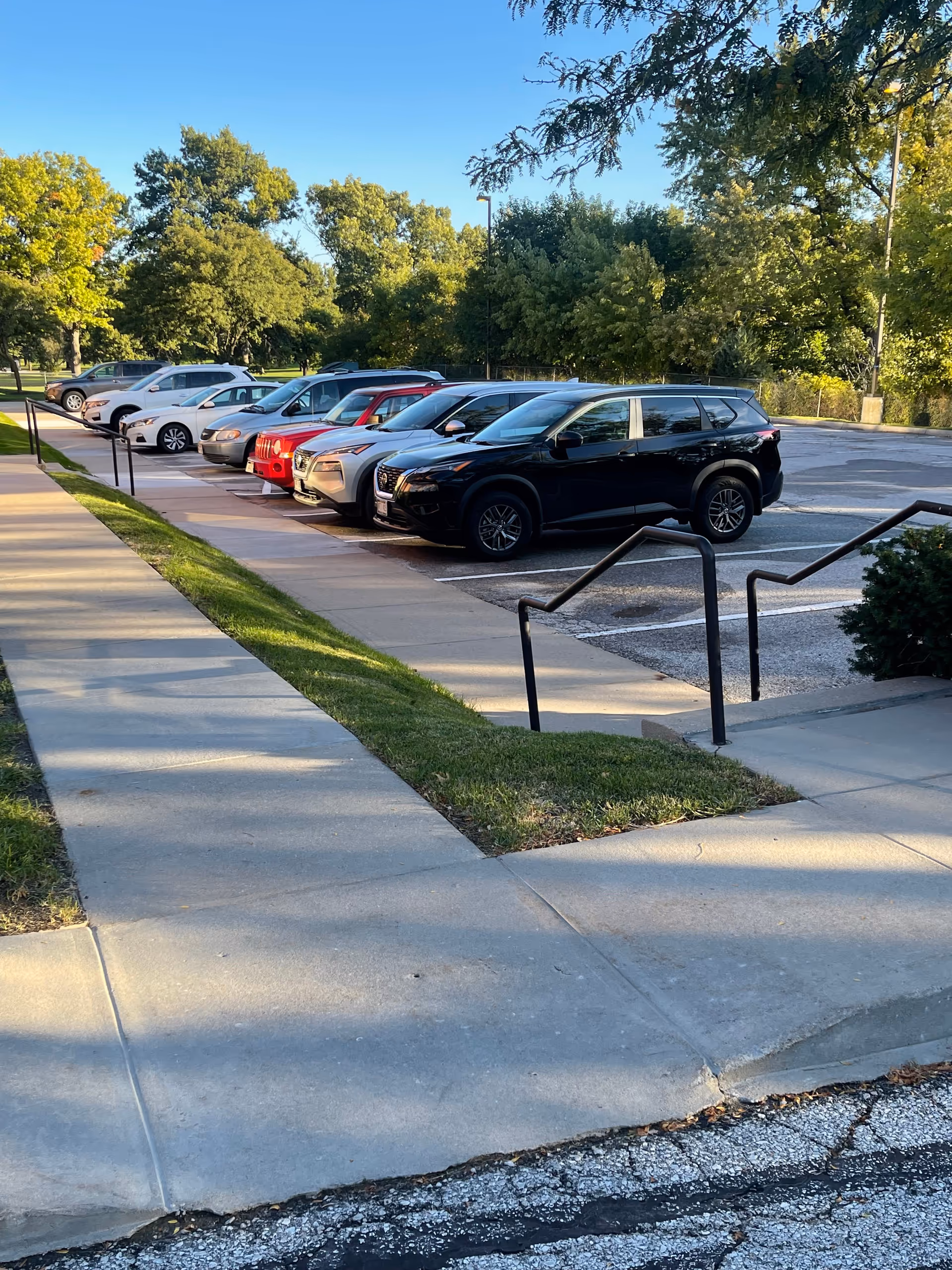 Exterior parking lot with several cars and surrounding trees beside a sidewalk.