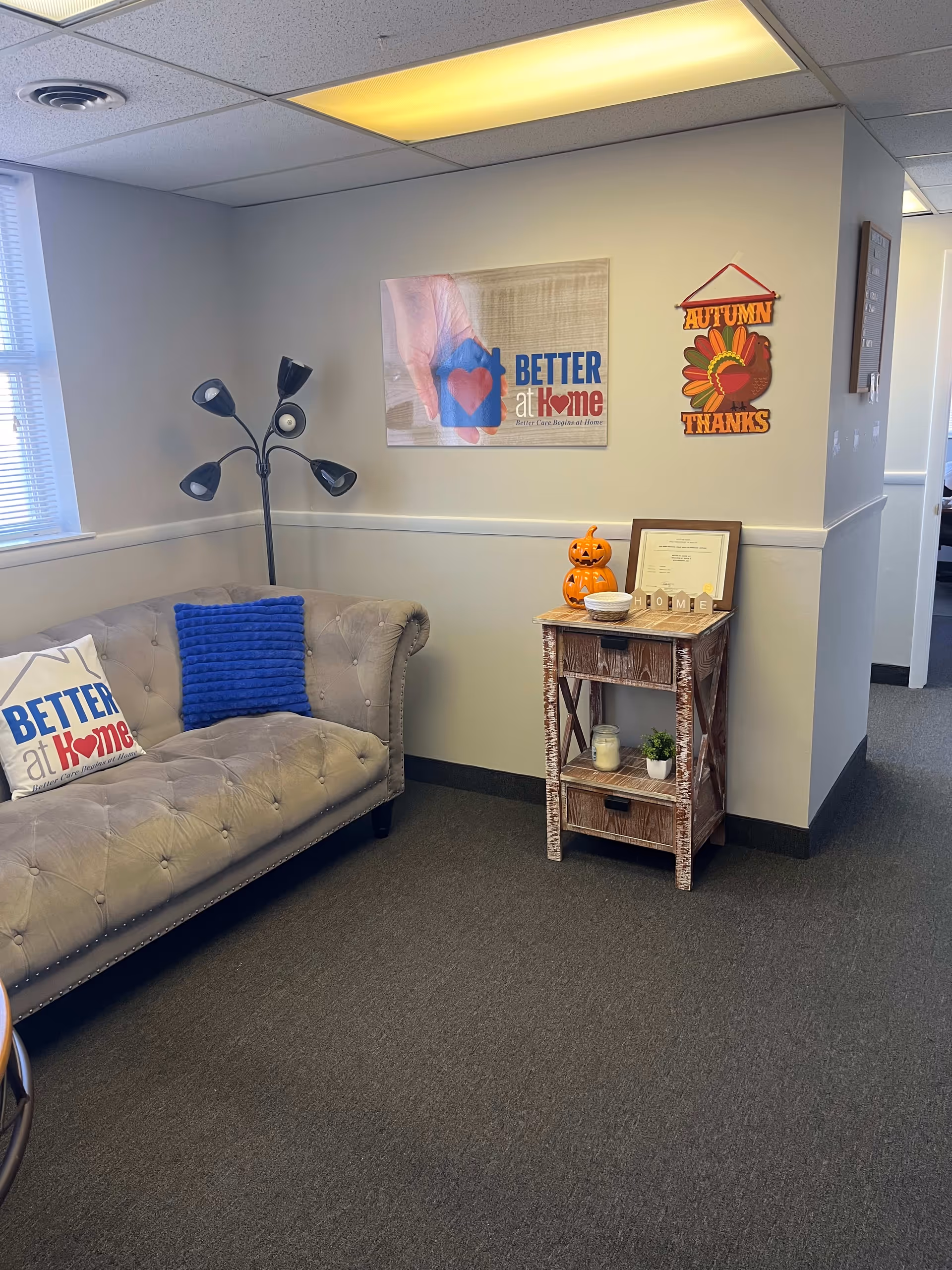 Cozy senior living lounge with tufted gray sofa, blue pillow, lamp, and autumn decor.