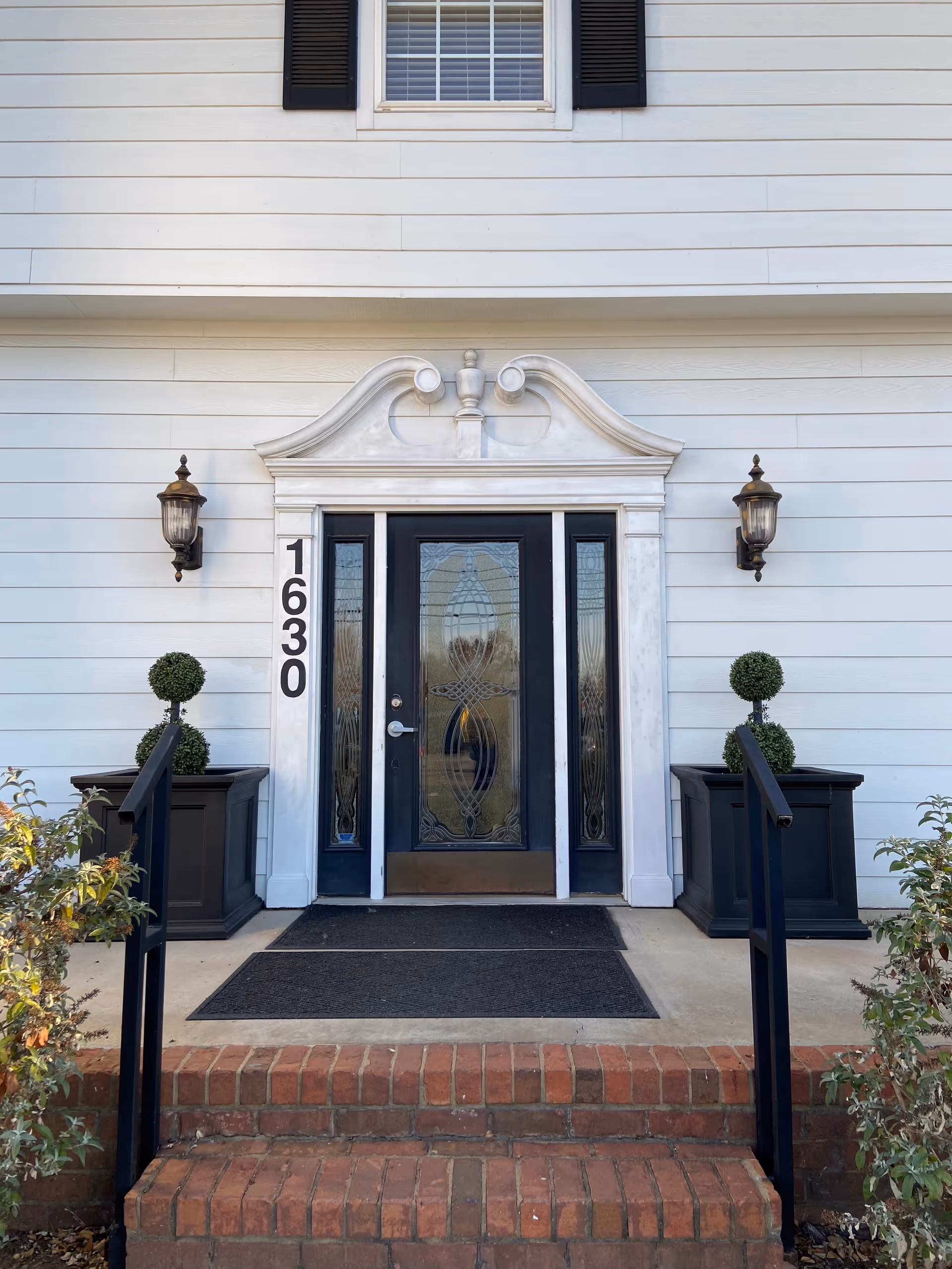 White building facade featuring an ornate doorway, black shutters, brick steps, and decorative planters.