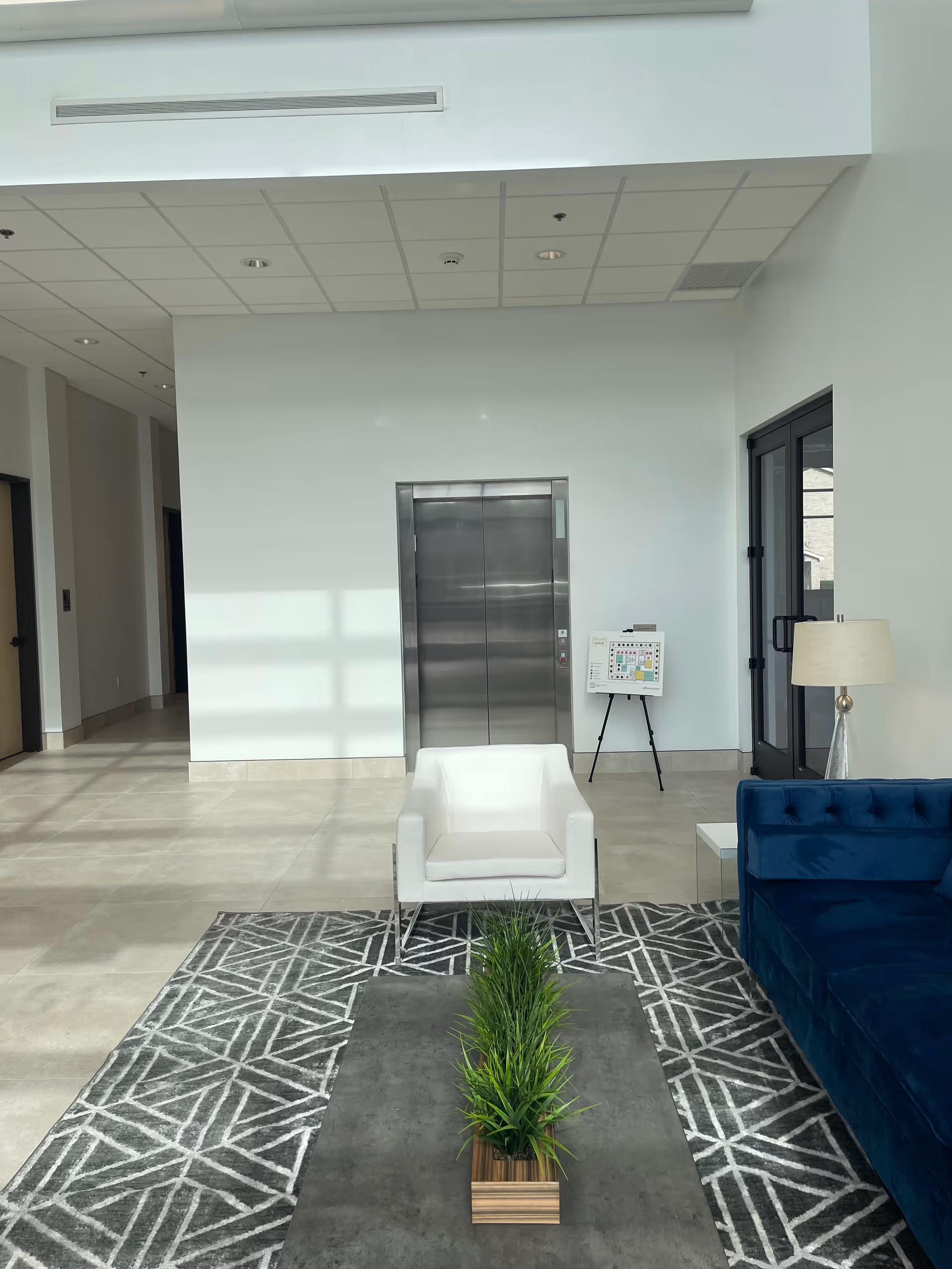 Modern, well-lit indoor lobby with elevator, white chair, blue couch, plants, and glass doors.