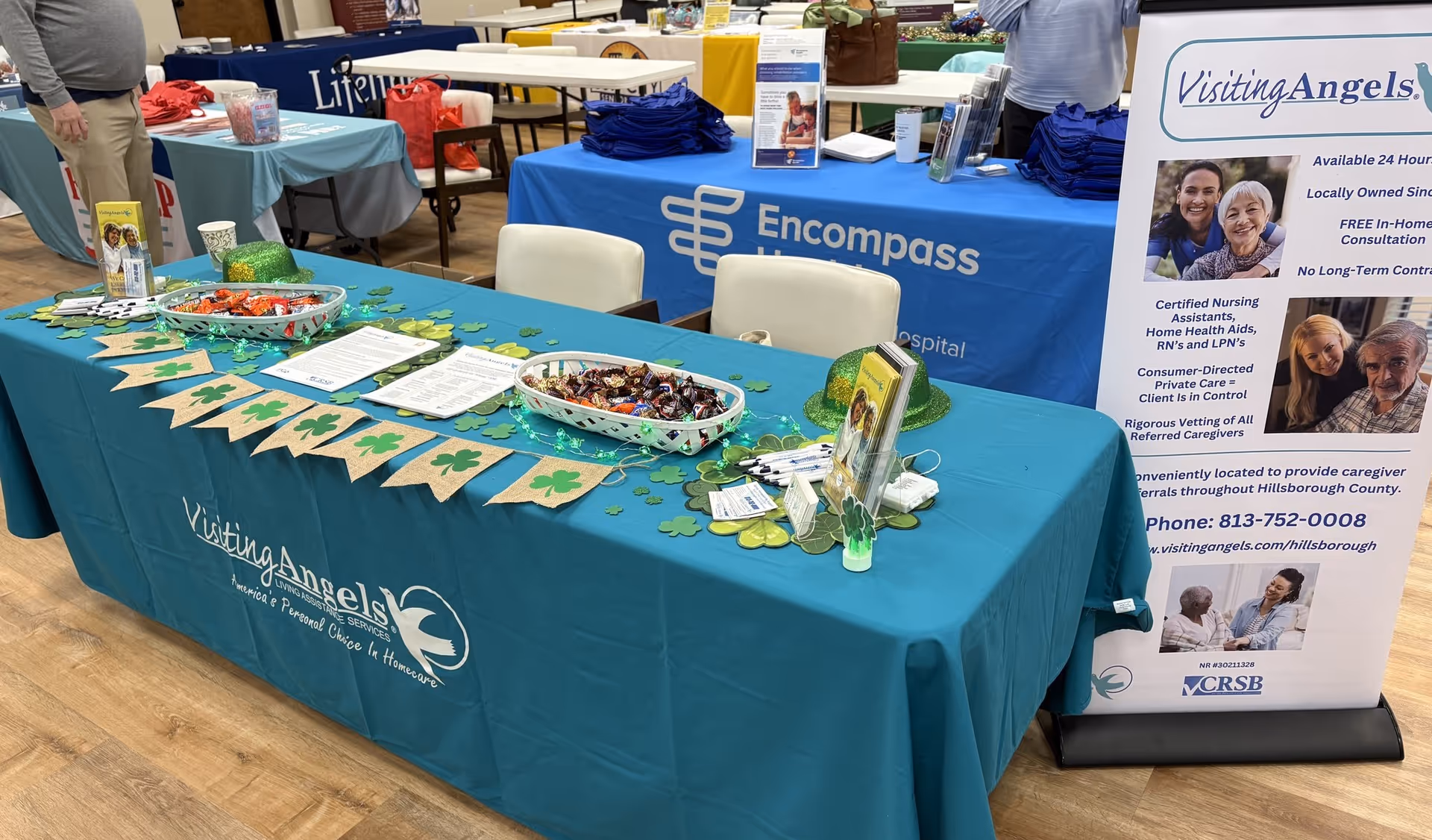 Indoor booth with teal tablecloths, banners, and cookies for Visiting Angels.