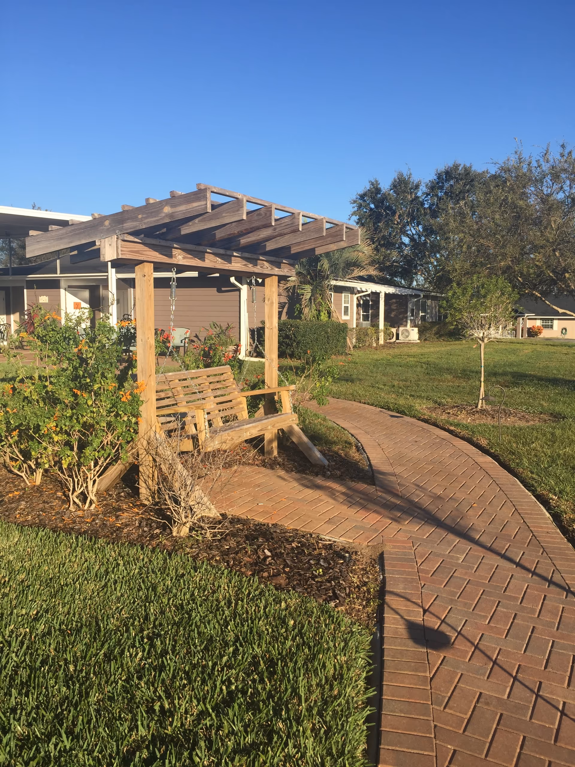Wood pergola with a bench swing beside a curved brick walkway in landscaped outdoor area.