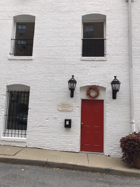 White brick building exterior with a red front door, wreath, and two lanterns.