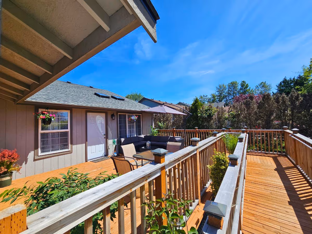 Outdoor wooden deck with furniture along a beige house under clear blue sky.