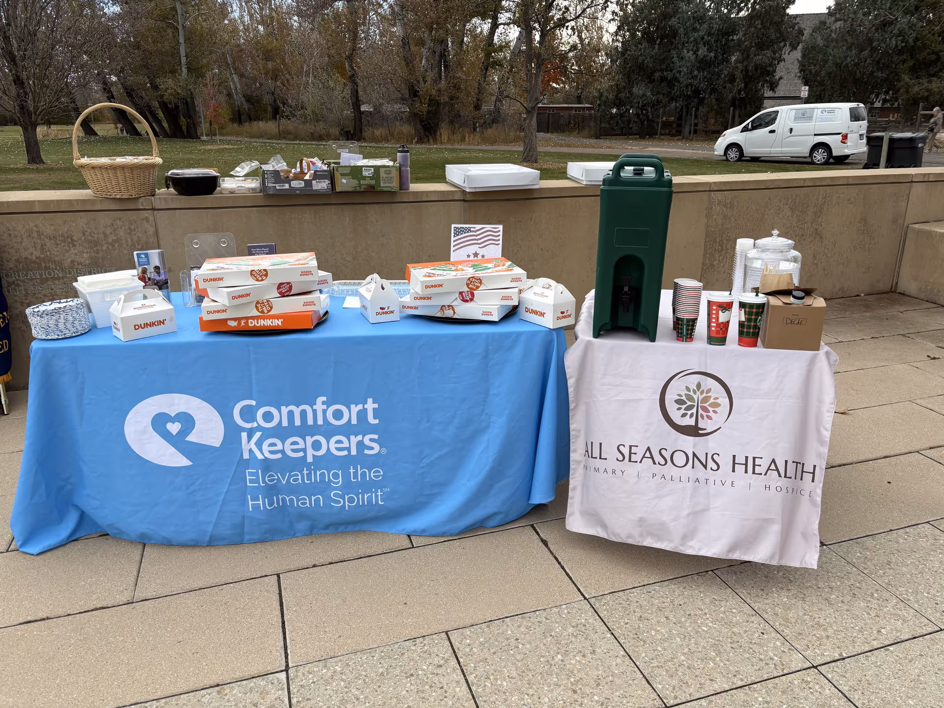 Two outdoor tables with Comfort Keepers and All Seasons Health banners, displaying food and beverages.