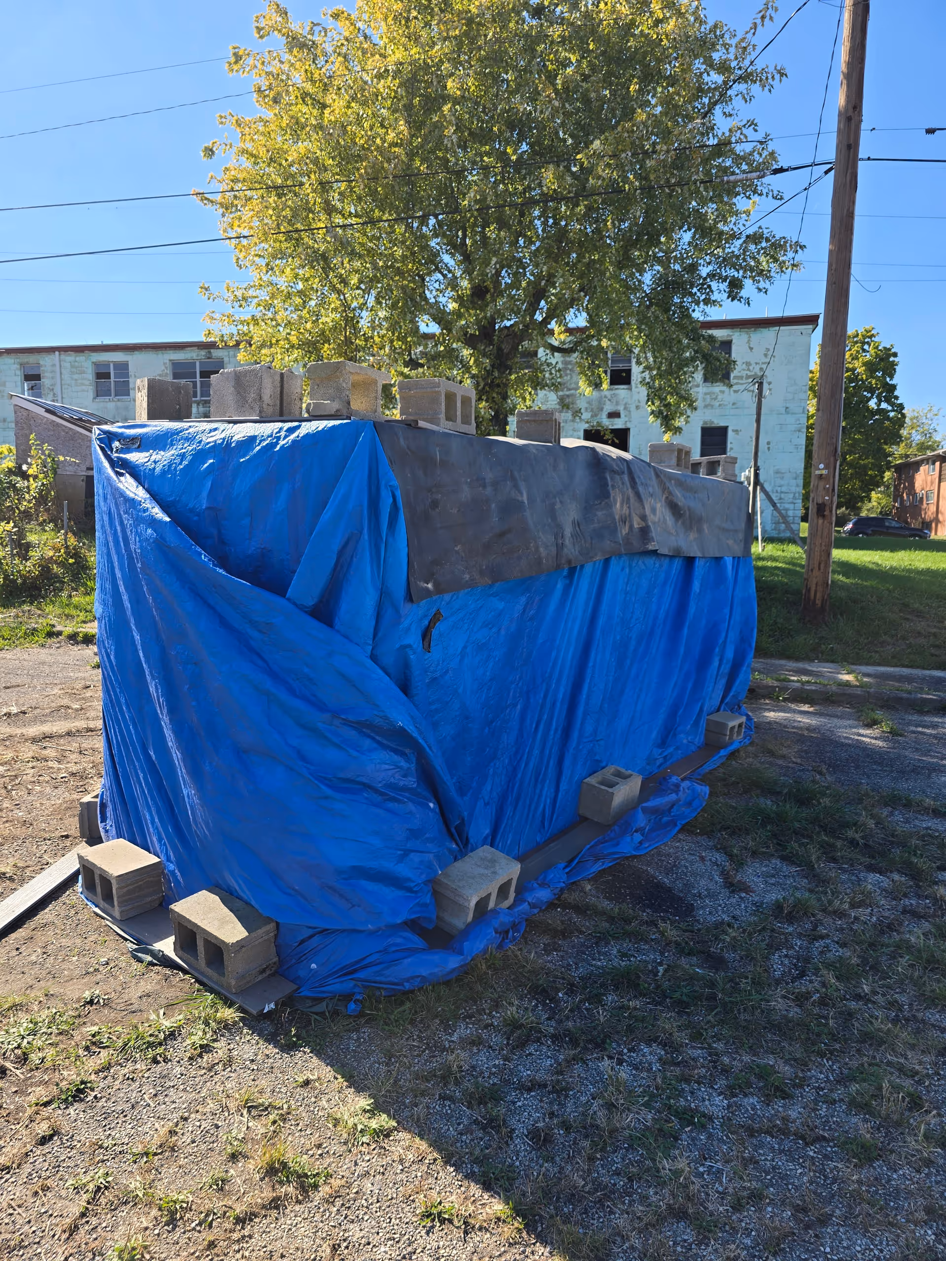 Blue tarp-covered container secured with concrete blocks sits in an outdoor lot near a tree.