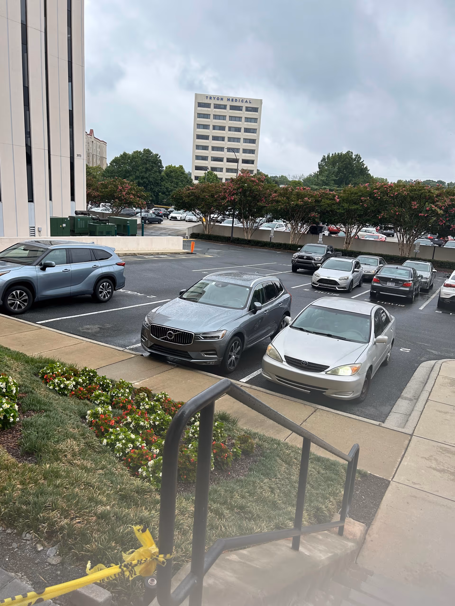 Outdoor parking lot with parked cars, railings, and flowering landscaping near a tall building.