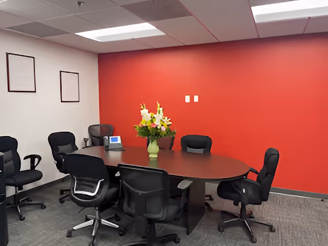 A conference room with a round table, black chairs, red wall, and flower centerpiece.