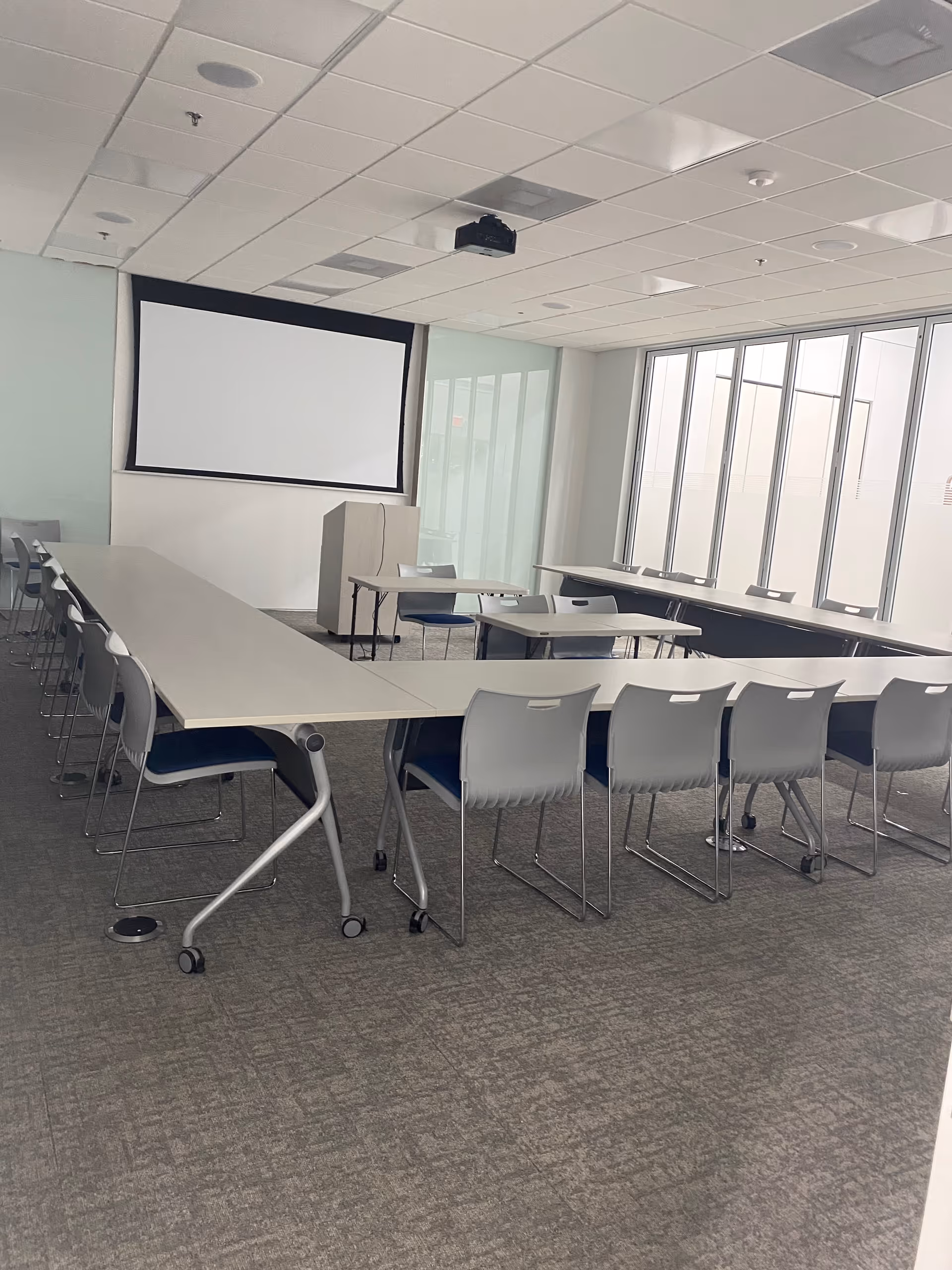 Empty conference room with long tables, chairs, and a projection screen at the front.