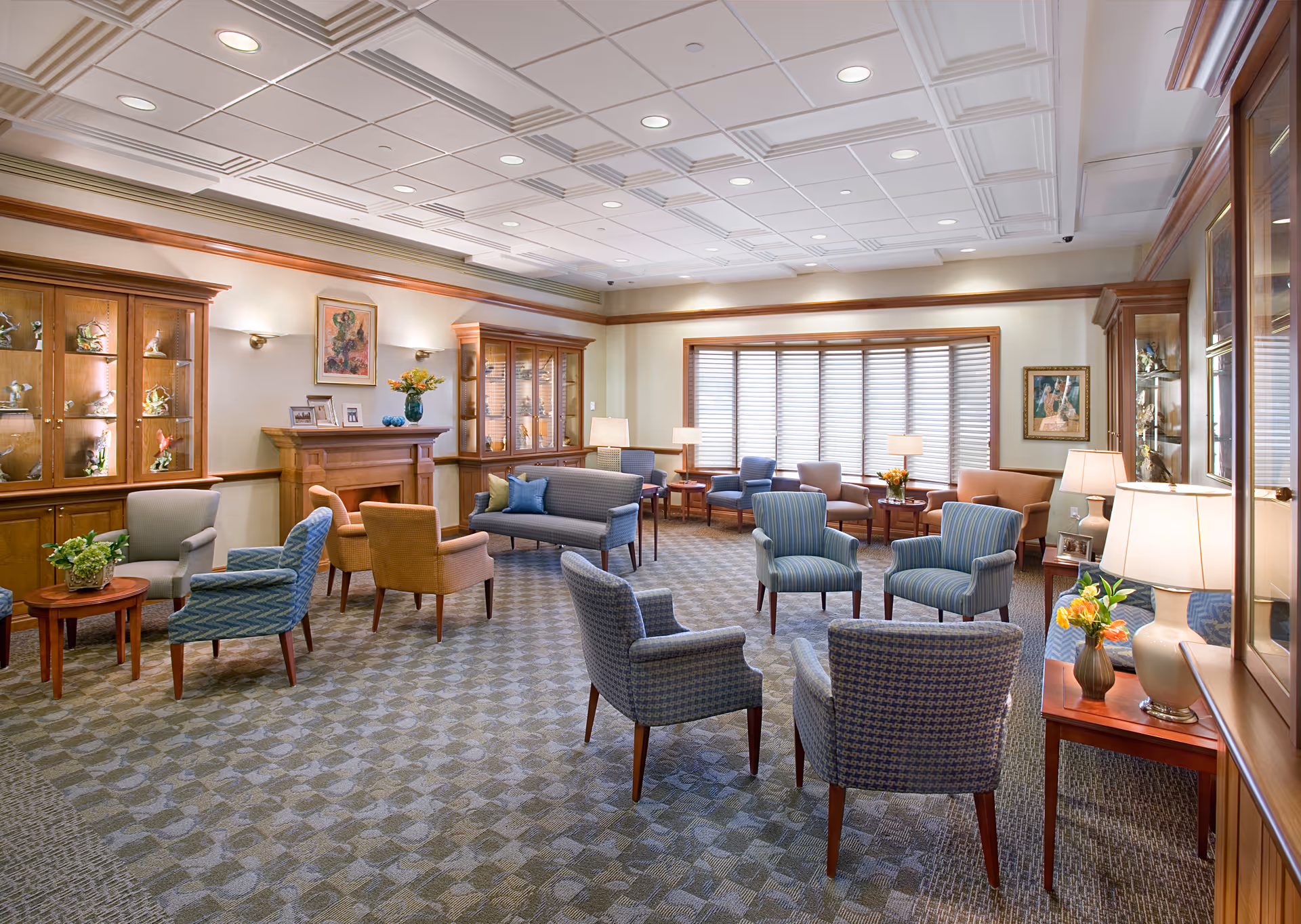 A spacious senior living lounge with chairs, tables, lamps, and display cabinets.