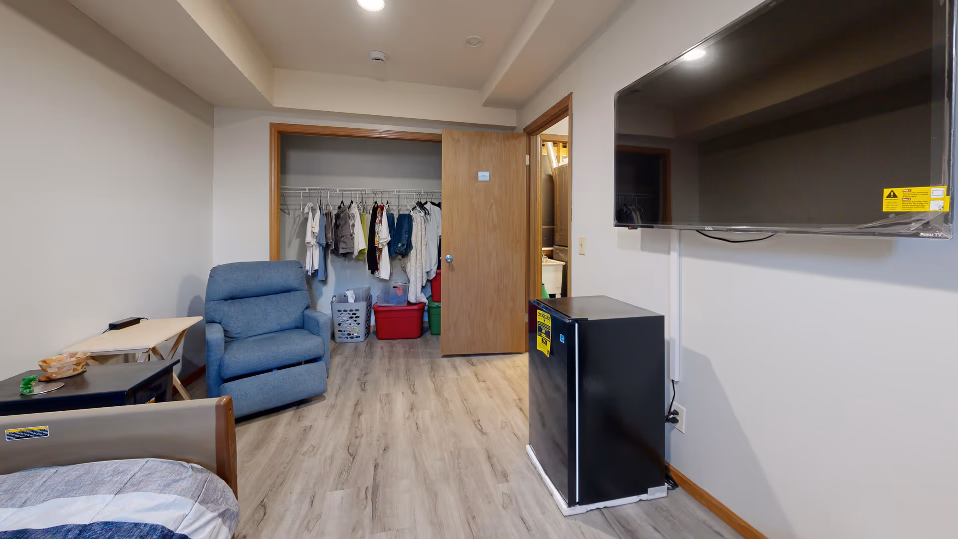 Small senior living suite with a bed, blue recliner, closet, TV, and mini-fridge.