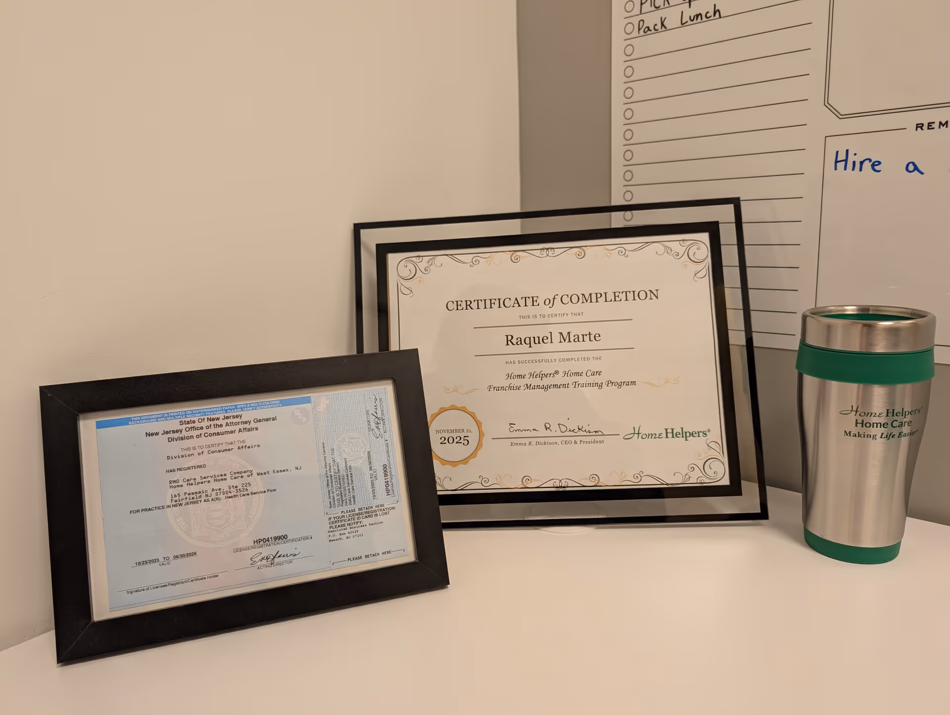 Framed certificates and a silver tumbler arranged on a desk against a white wall.