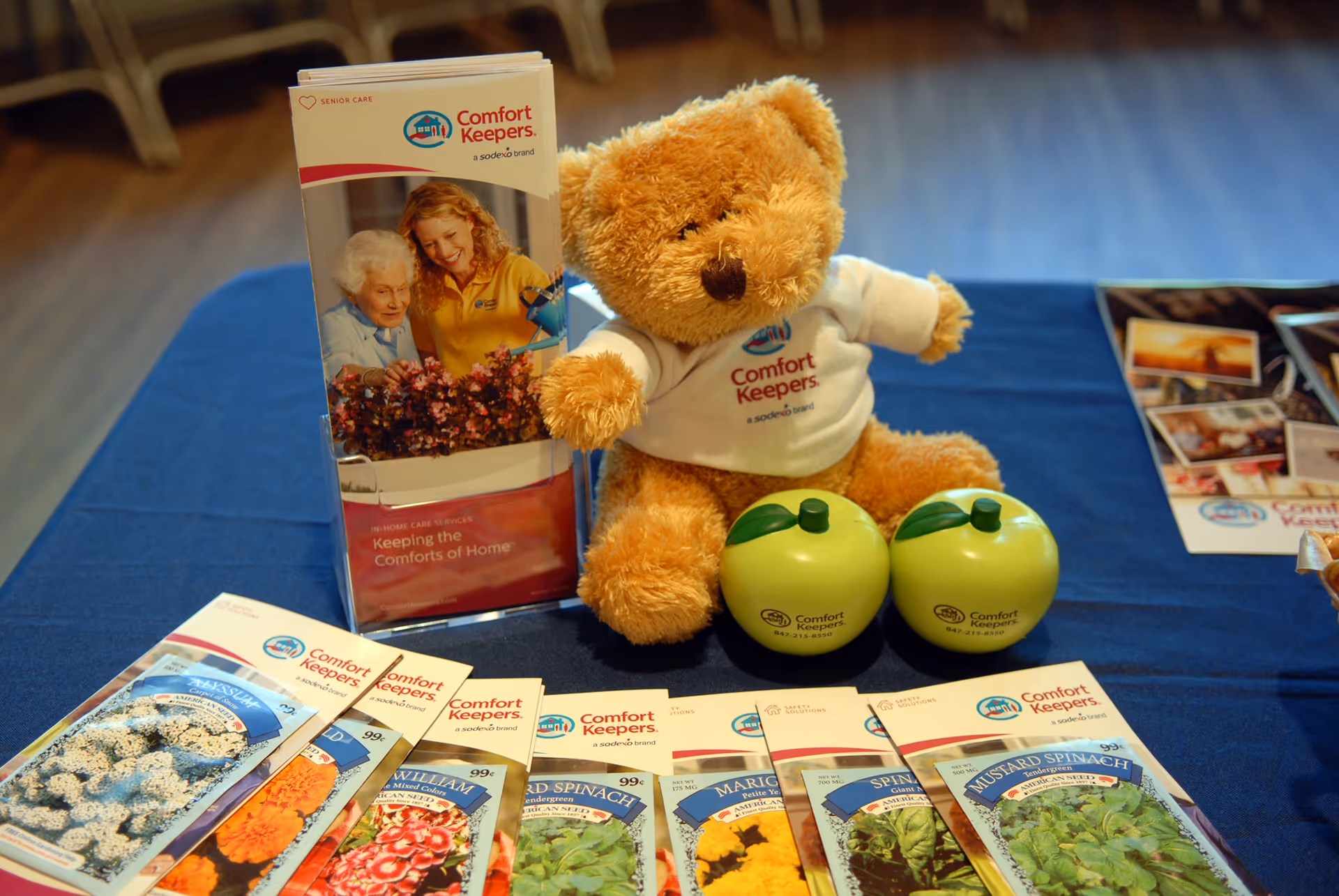 A teddy bear in a Comfort Keepers shirt sits beside brochures and two green apples.
