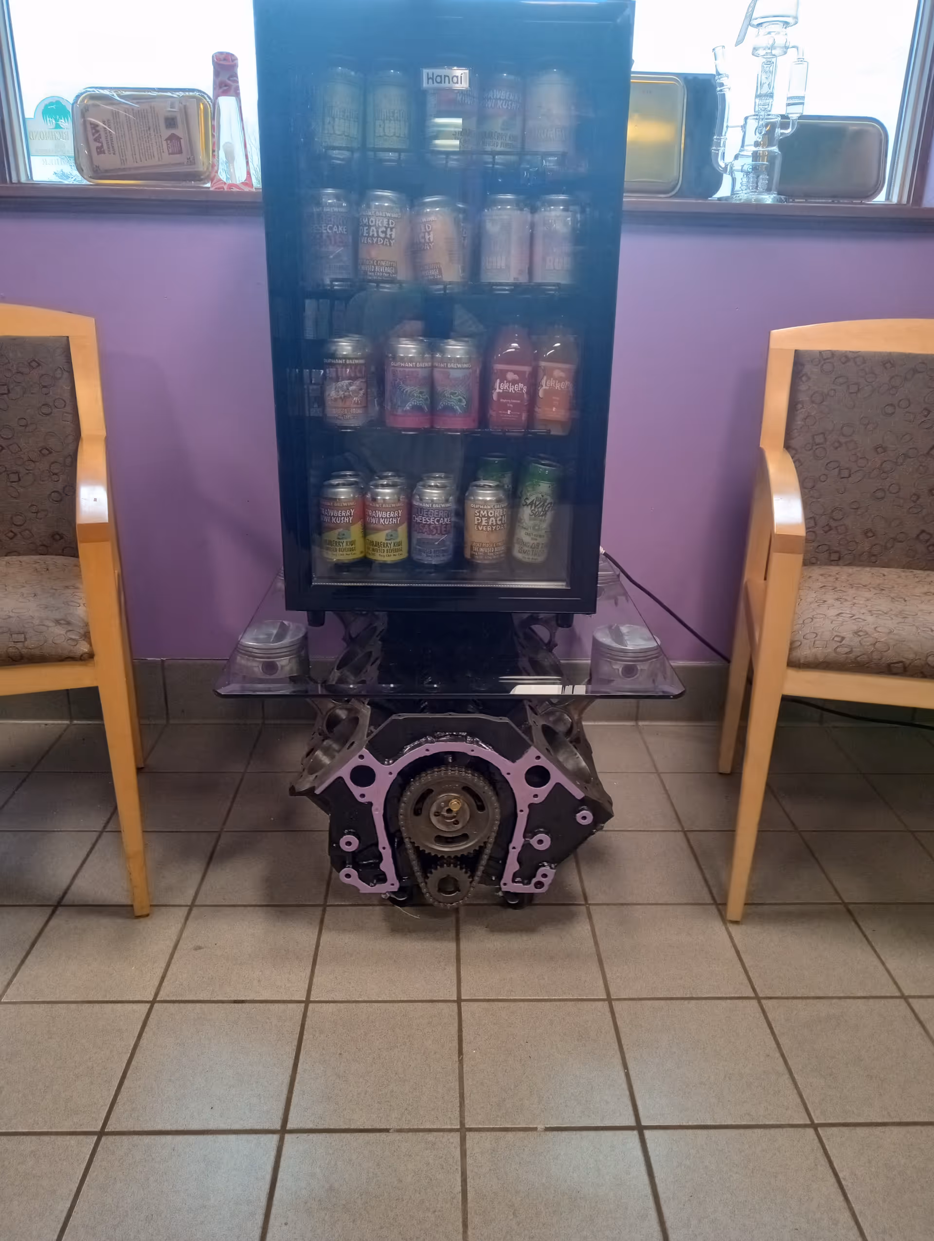 A beverage vending fridge sits on an engine-like base between two chairs in a purple interior.