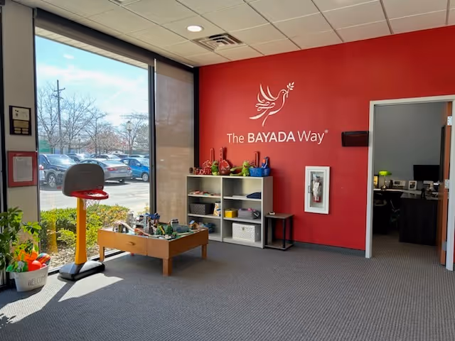 Bright interior lobby with red branding wall reading The BAYADA Way and children's play area.
