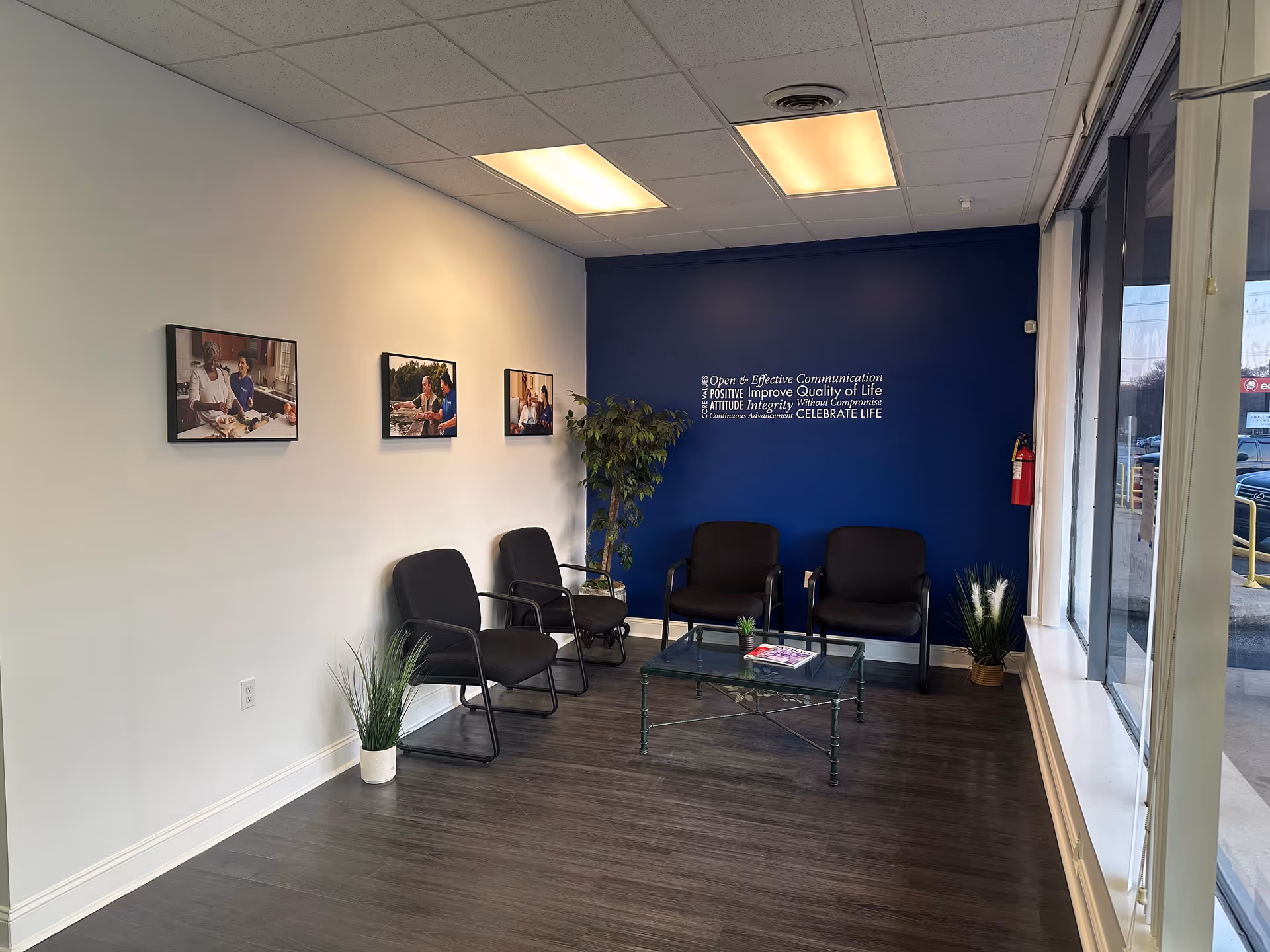 Waiting area with black chairs, glass coffee table, blue accent wall, photos, plants.