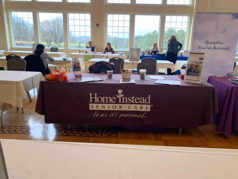 Indoor event hall with a long Home Instead-branded table and several attendees.