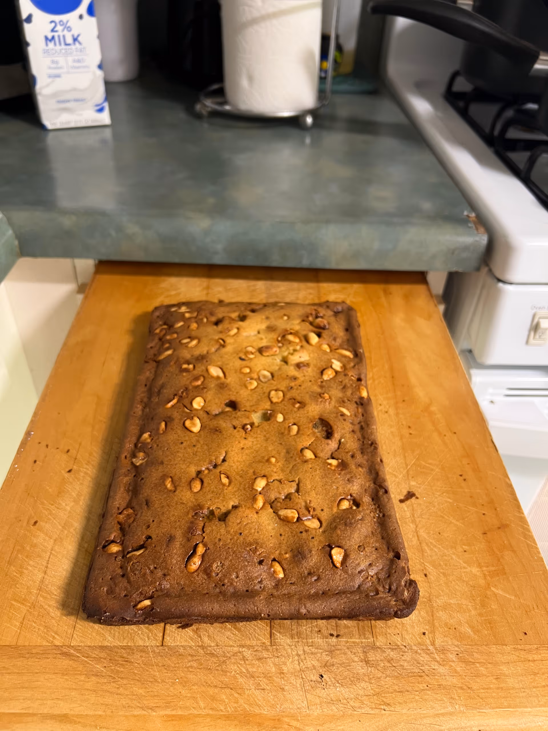 A loaf of nut-topped banana bread sits on a wooden cutting board in a kitchen.