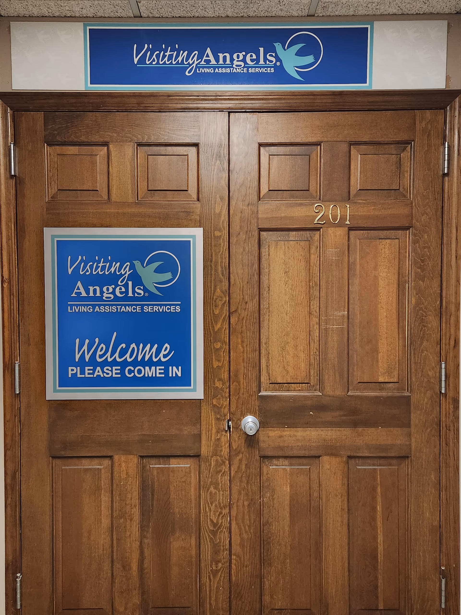Double wooden doors with blue Visiting Angels signs and a welcome poster.
