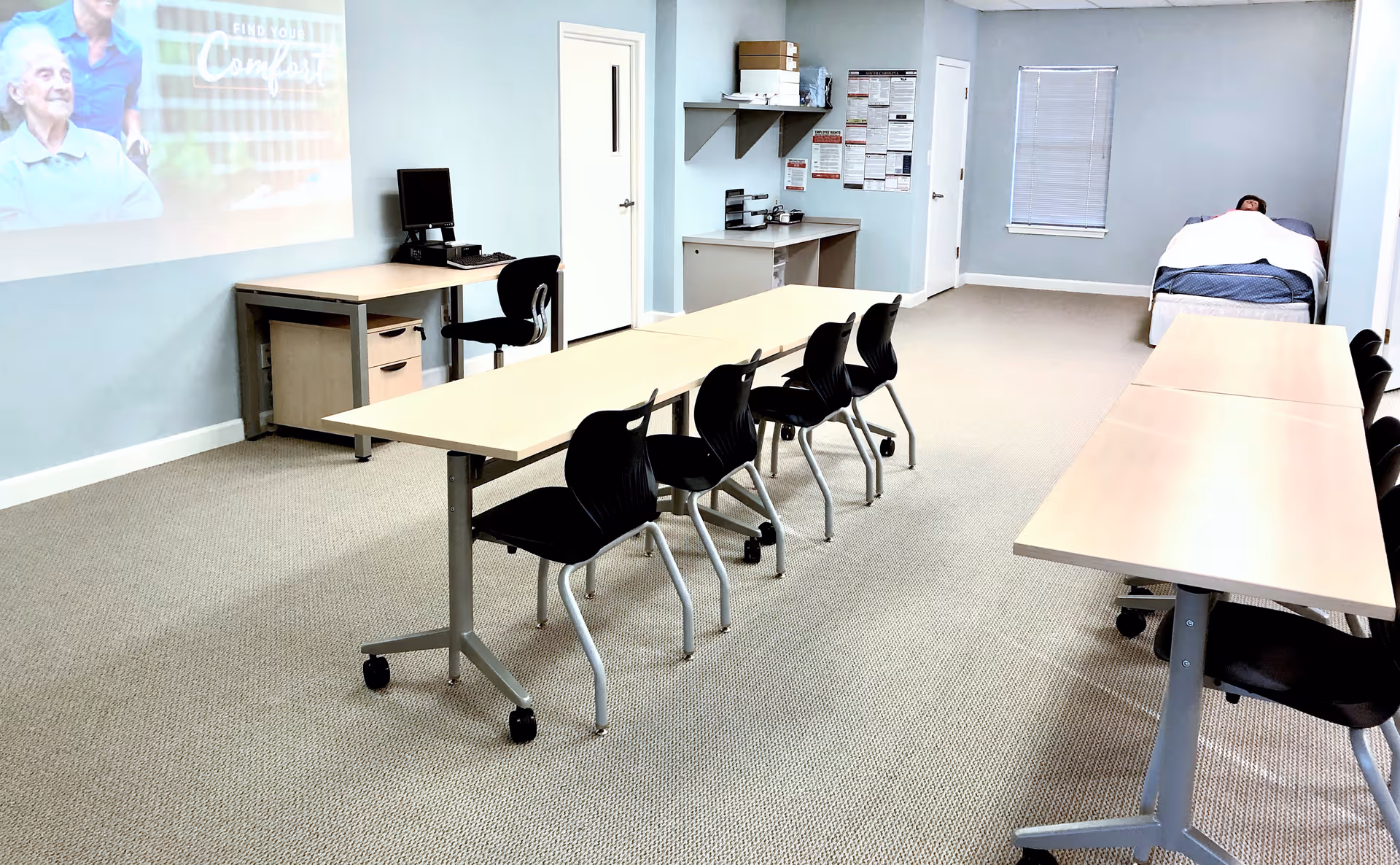 Light blue interior room with rows of tables, chairs, projector, and a bed at the back.