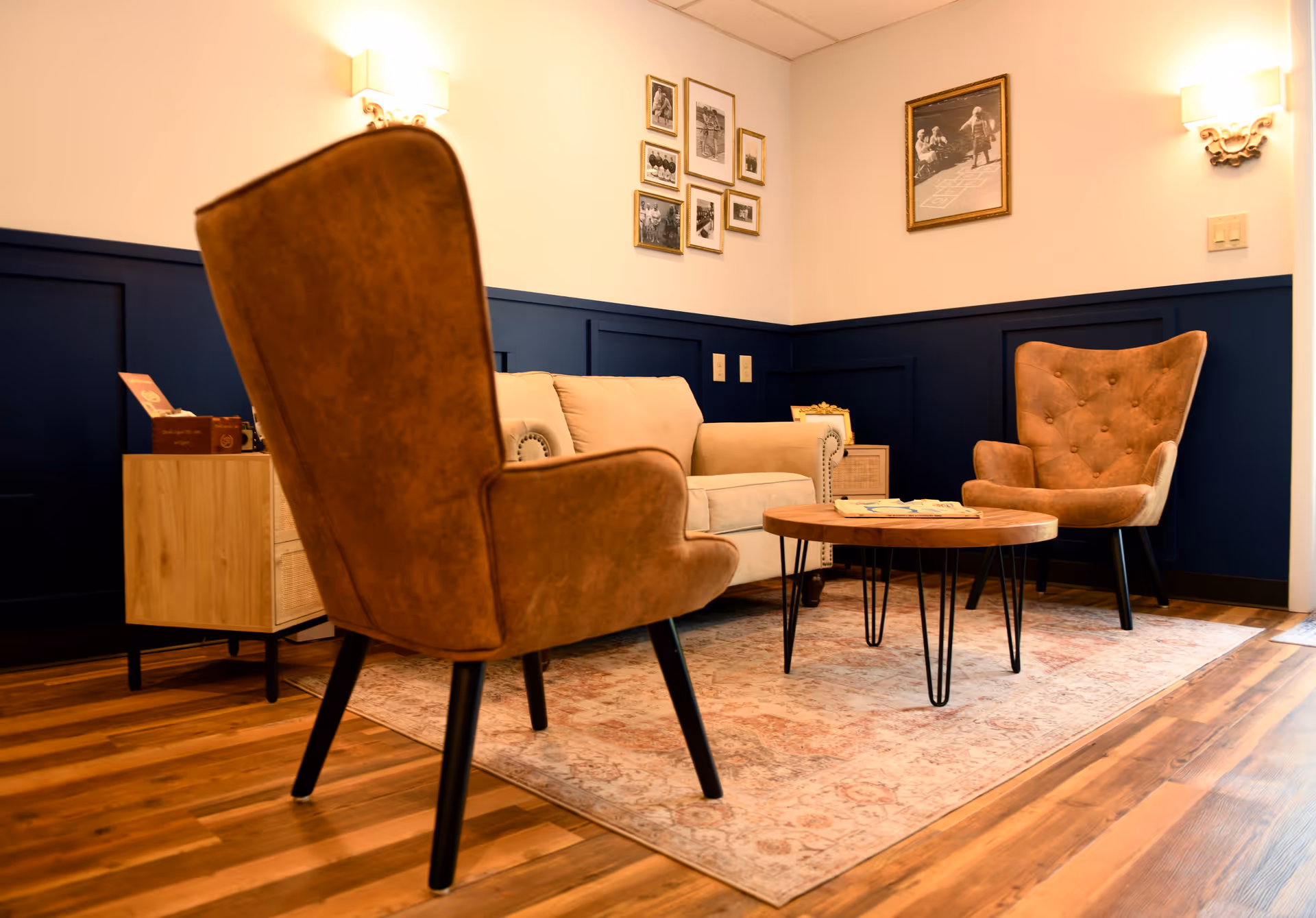 Cozy senior living lounge with tan upholstered chairs, sofa, round coffee table, rug, and framed photos.