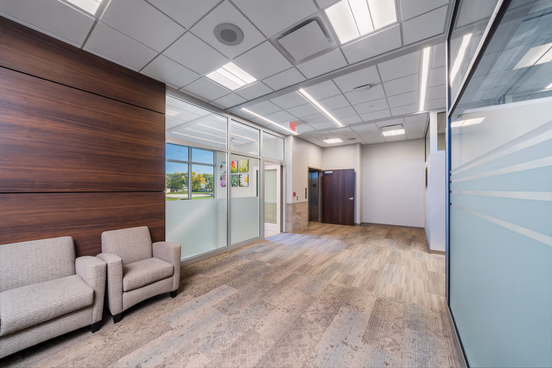 Interior hallway of a medical facility with frosted glass walls, wood paneling, and beige lounge chairs.