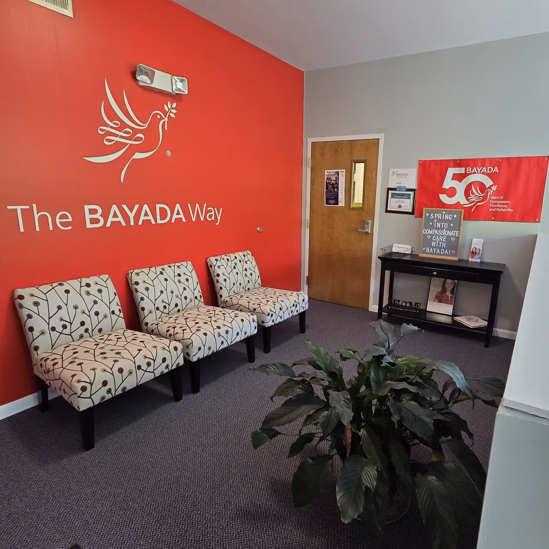 Lobby area with red BAYADA wall, The BAYADA Way text, four patterned chairs, doorway, plant.