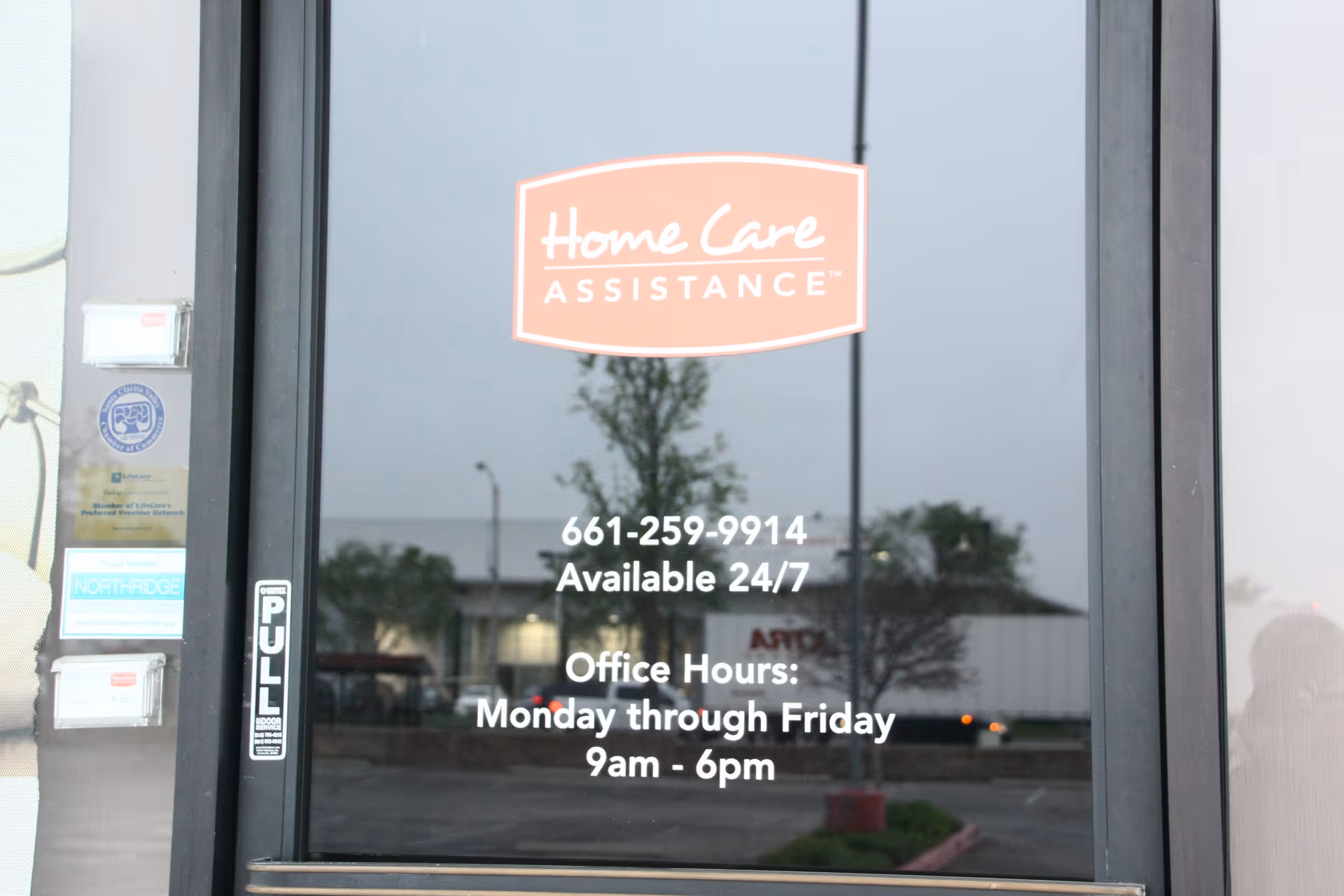 Glass door displays Home Care Assistance logo and office hours, reflecting a building exterior.