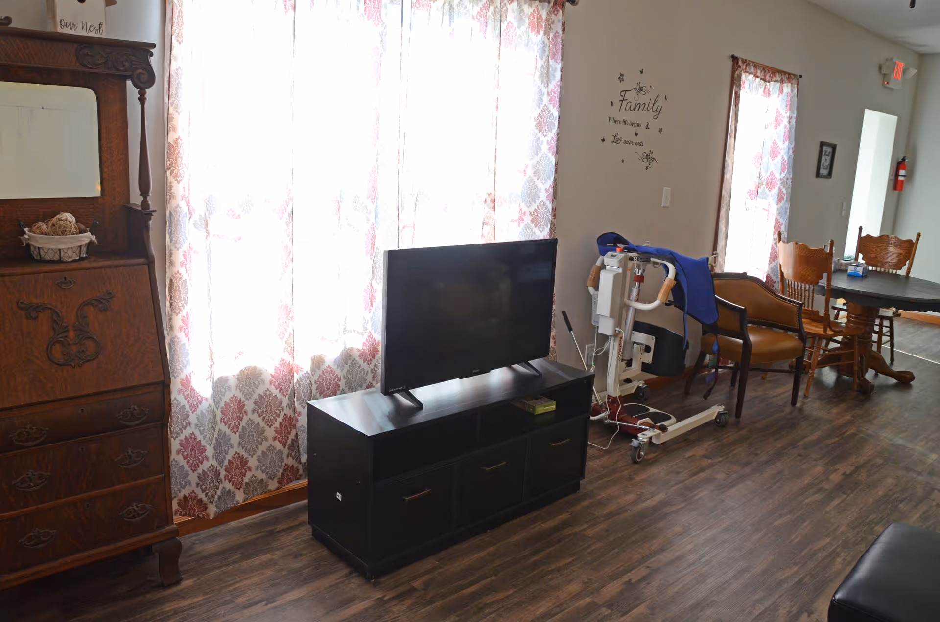 TV on stand in a living room with patterned curtains, chairs, and a patient lift.