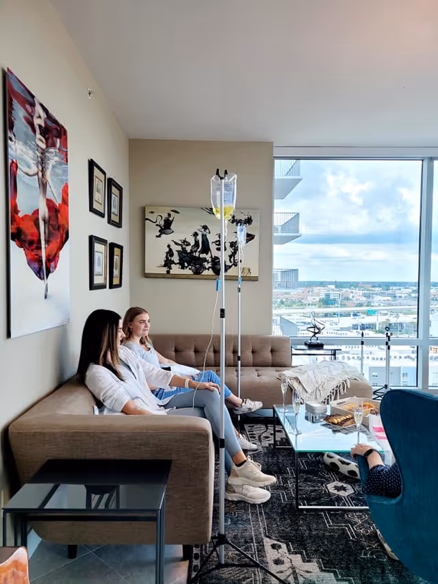 Two residents sit on a beige sofa in a bright living room with IV stands.