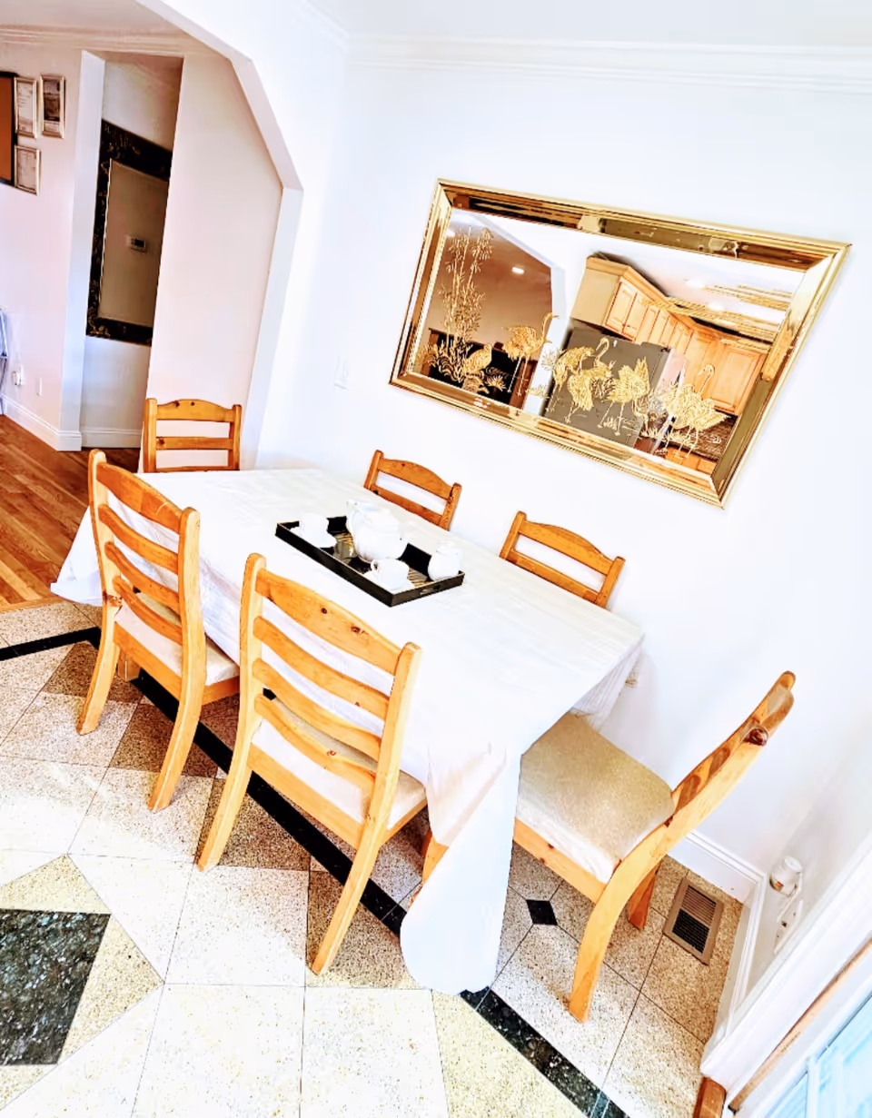 A dining room with a long table, six wooden chairs, and a large gold-framed wall mirror.