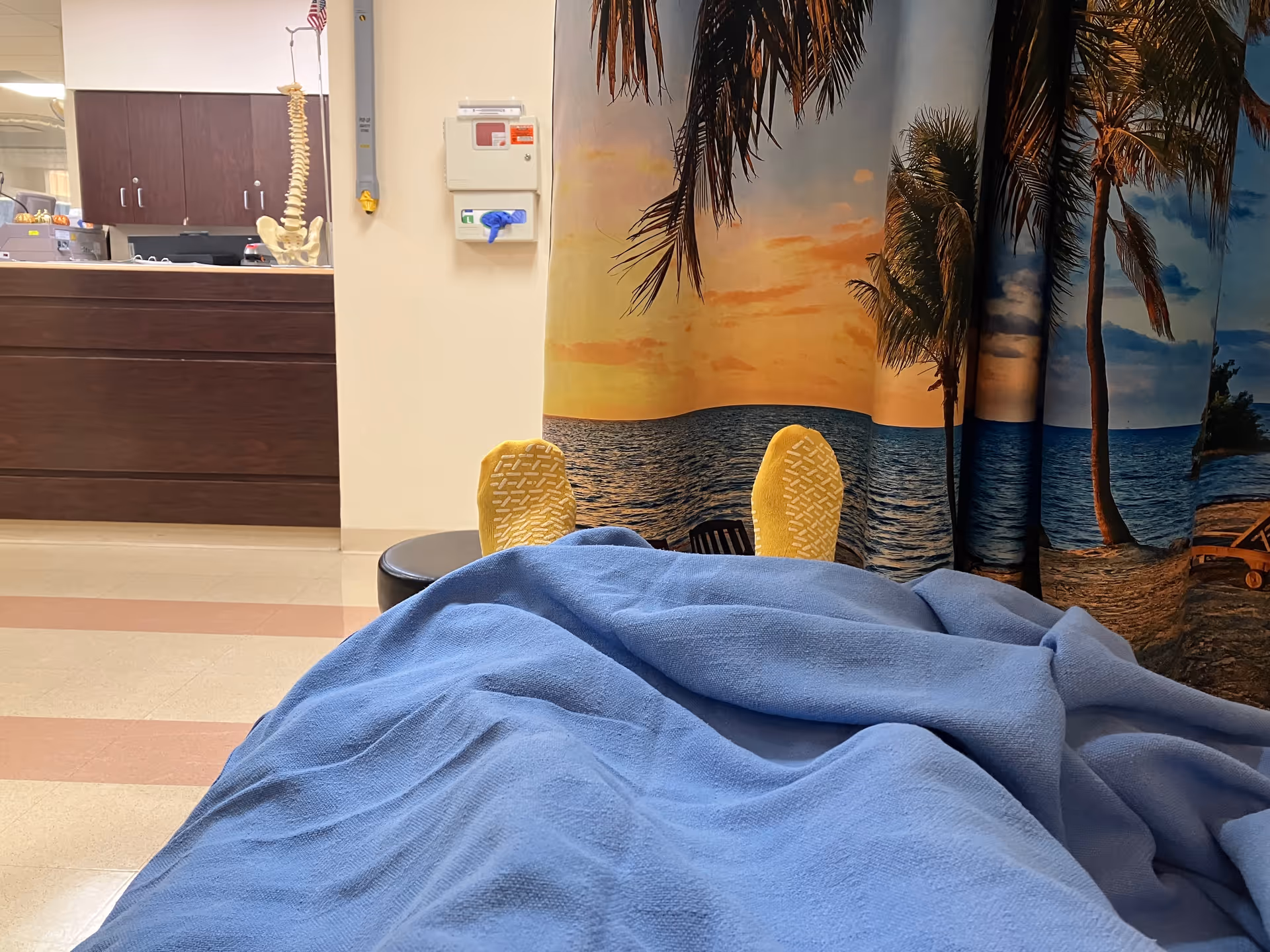 Blue blanket covers a hospital bed in front of a tropical beach mural.