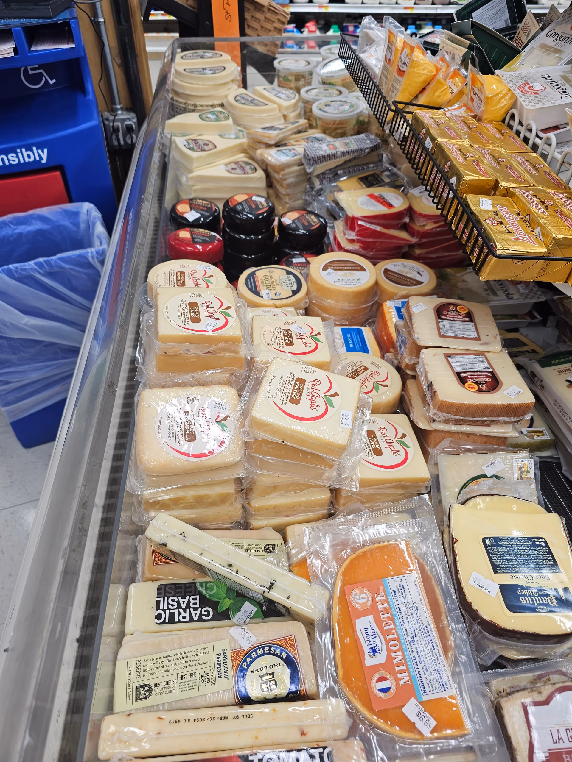Refrigerated dairy aisle with assorted cheeses in clear plastic packaging.