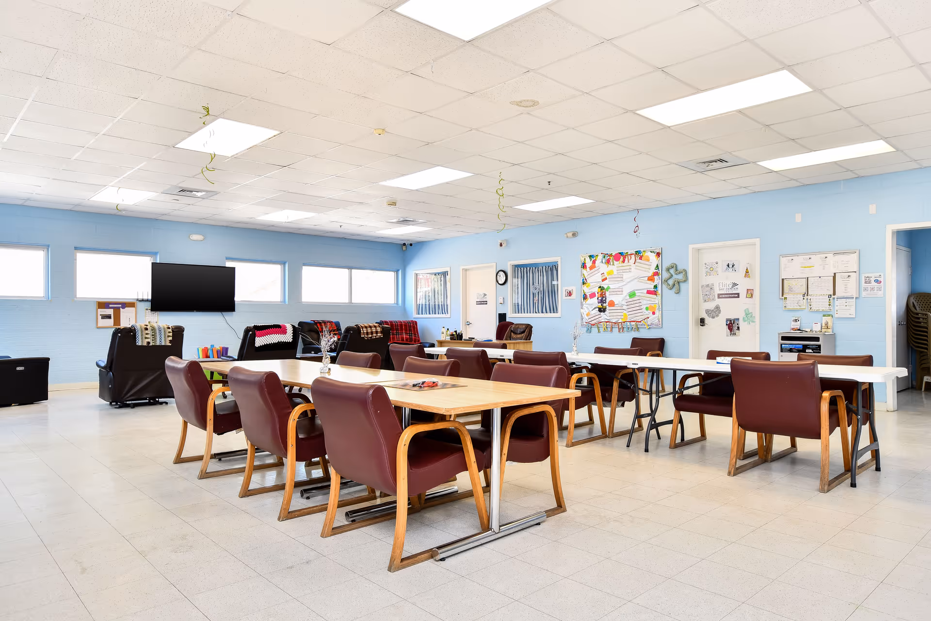 Bright senior living common room with multiple tables, burgundy chairs, and a wall of windows.