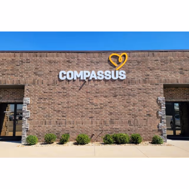 Brick facility exterior with Compassus sign, heart logo, glass doors, and landscaped shrubs.