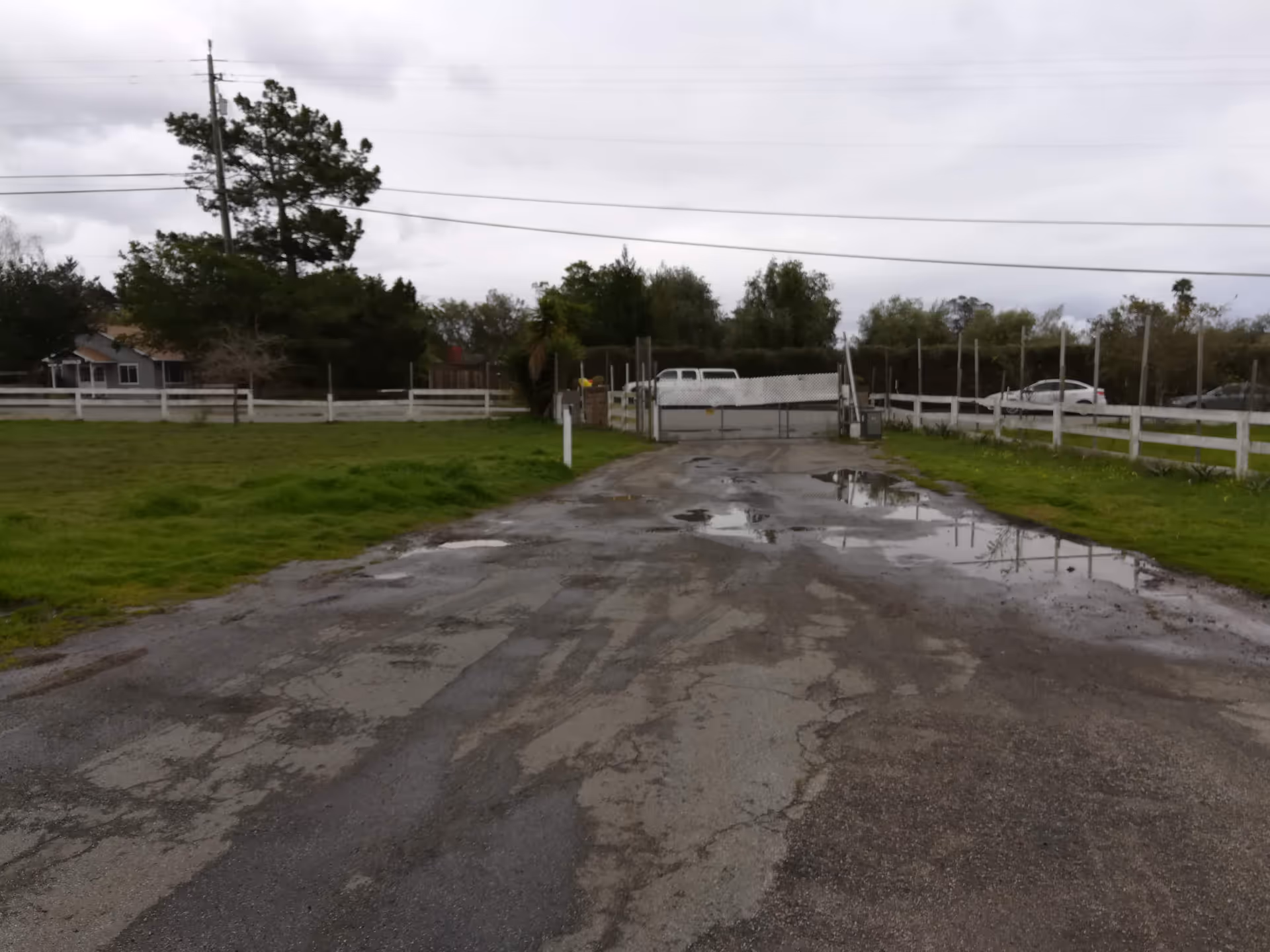 Paved driveway with puddles leads to a gated facility entrance, surrounded by grassy fields.