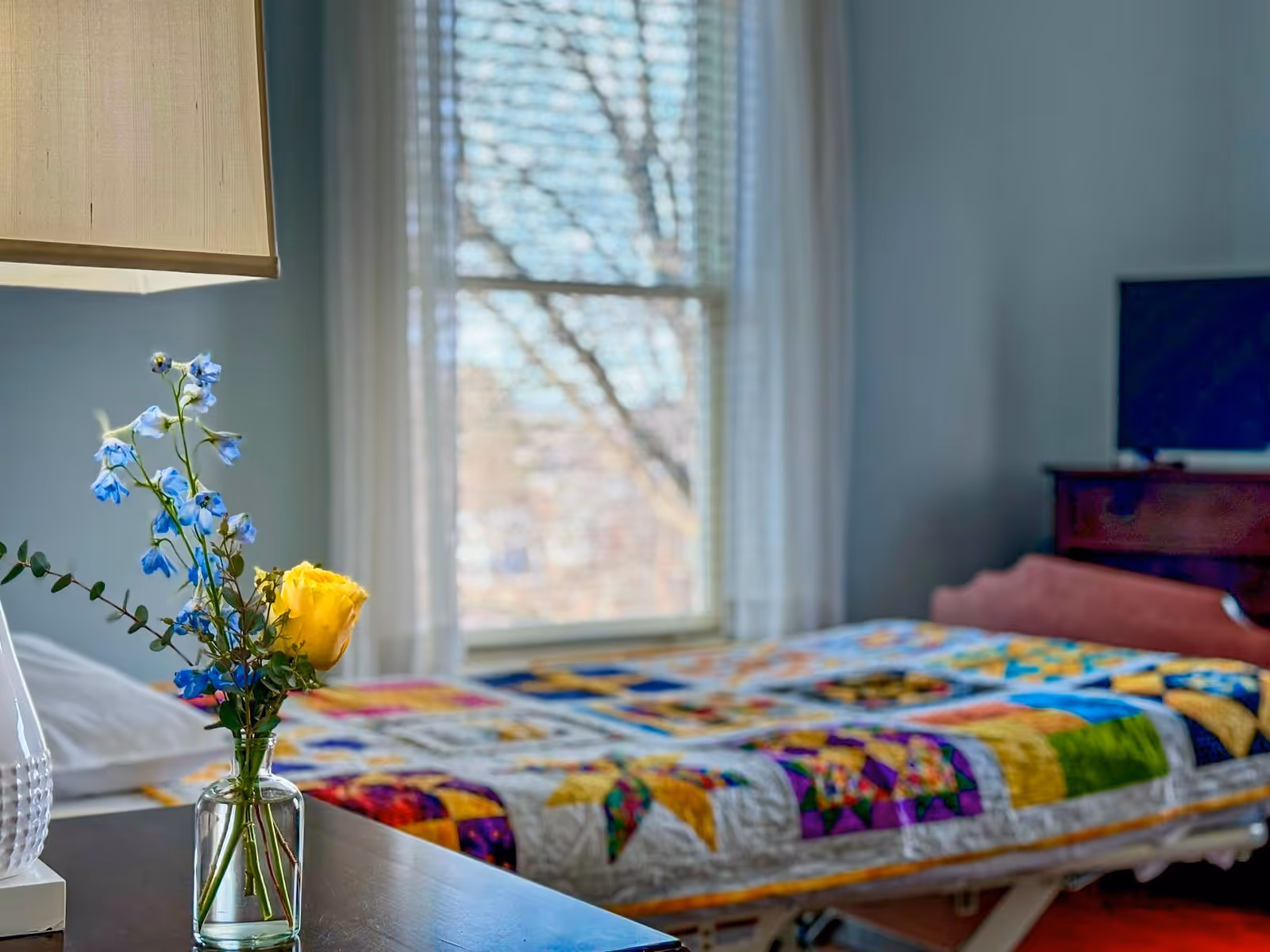 A cozy bedroom with a colorful quilt, flowers on a nightstand, and a window.