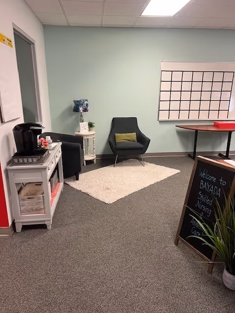 Cozy indoor lounge with a chair, rug, small table, lamp, and a welcoming chalkboard sign.