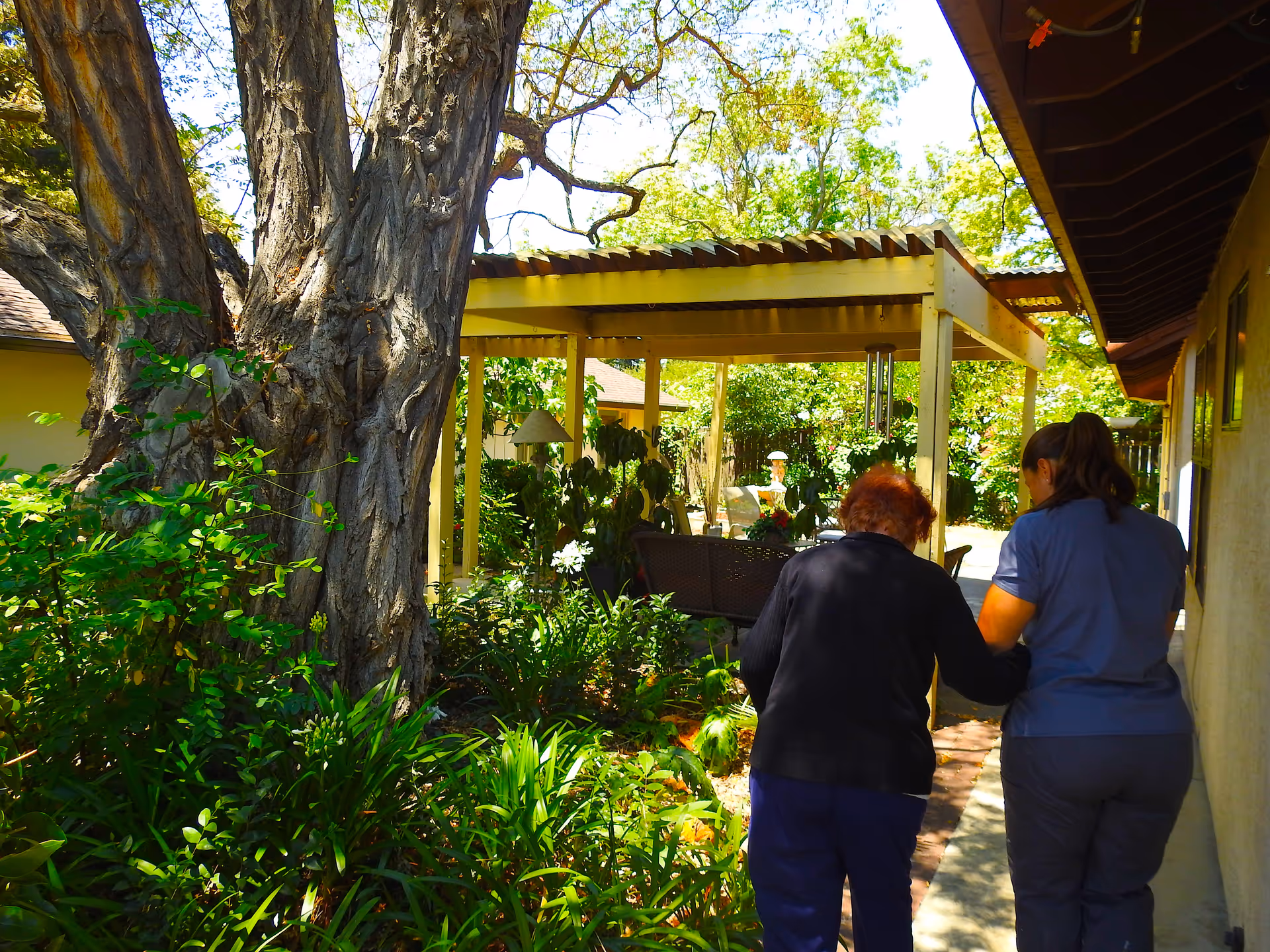 Two caregivers assist an elderly resident through a shaded outdoor garden area.