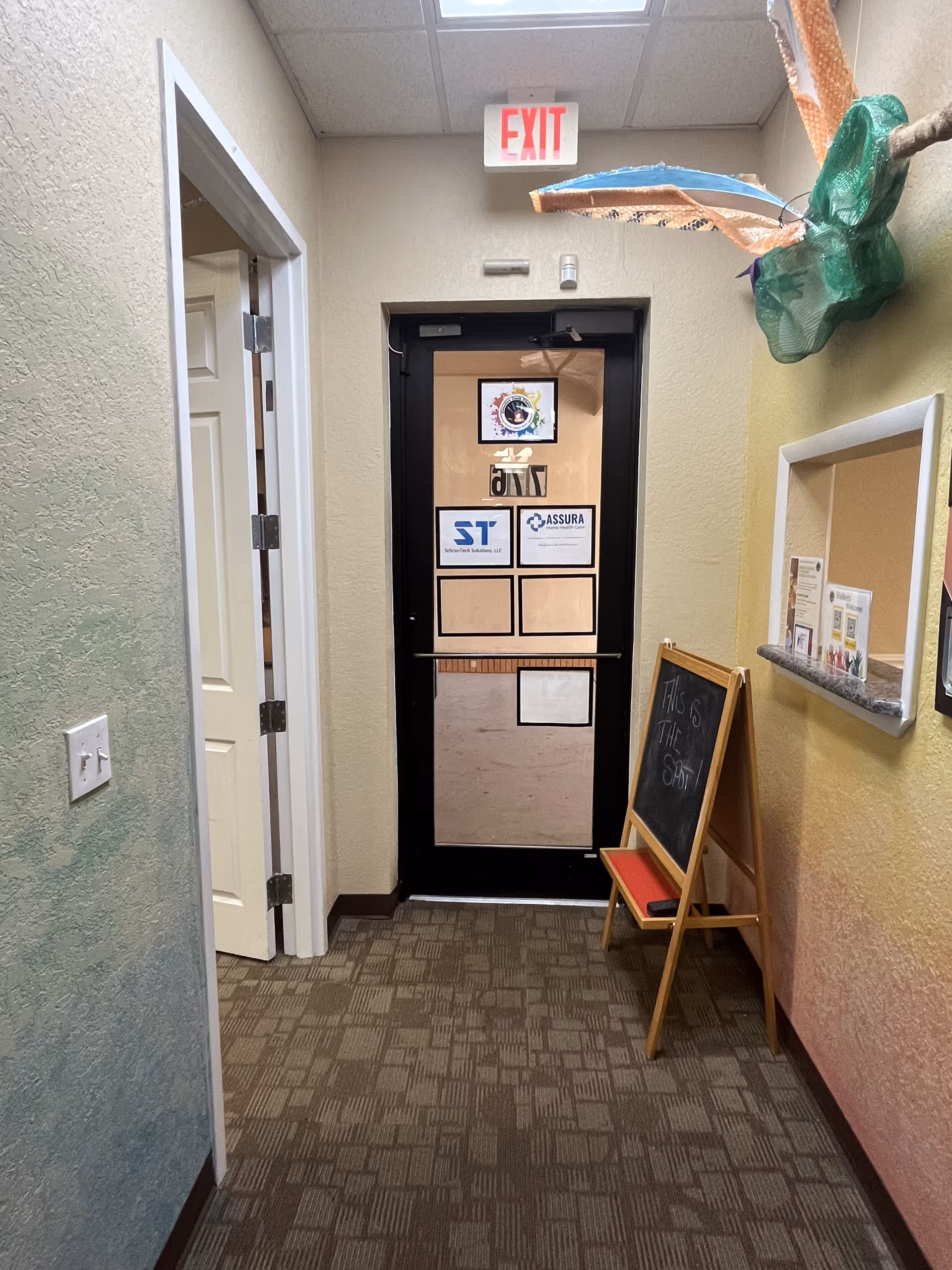 Interior hallway of a senior facility with an exit door, bulletin board, and a chalkboard stand.