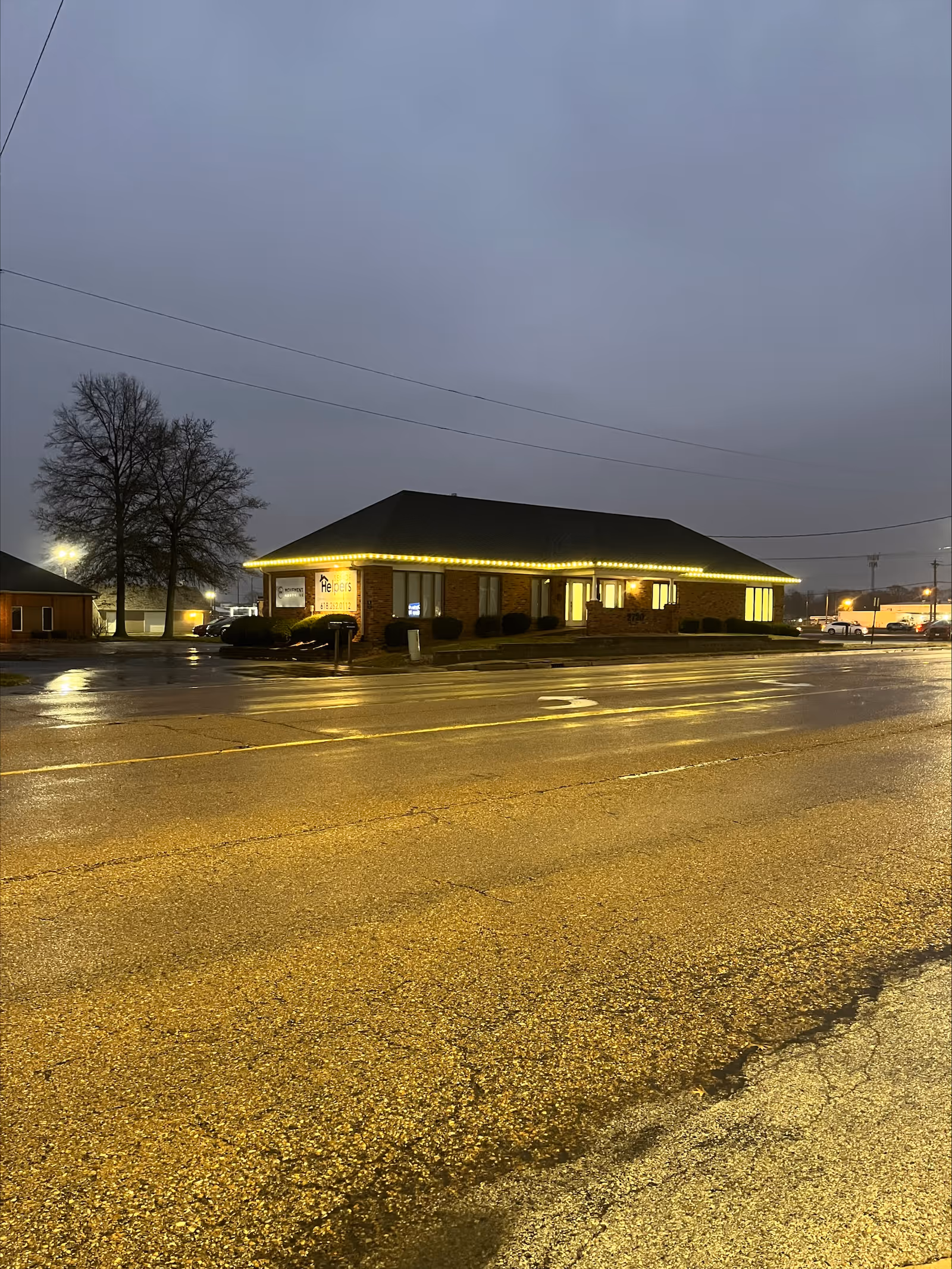 Exterior view of a Senior Helpers facility at dusk with illuminated signage along a wet street.