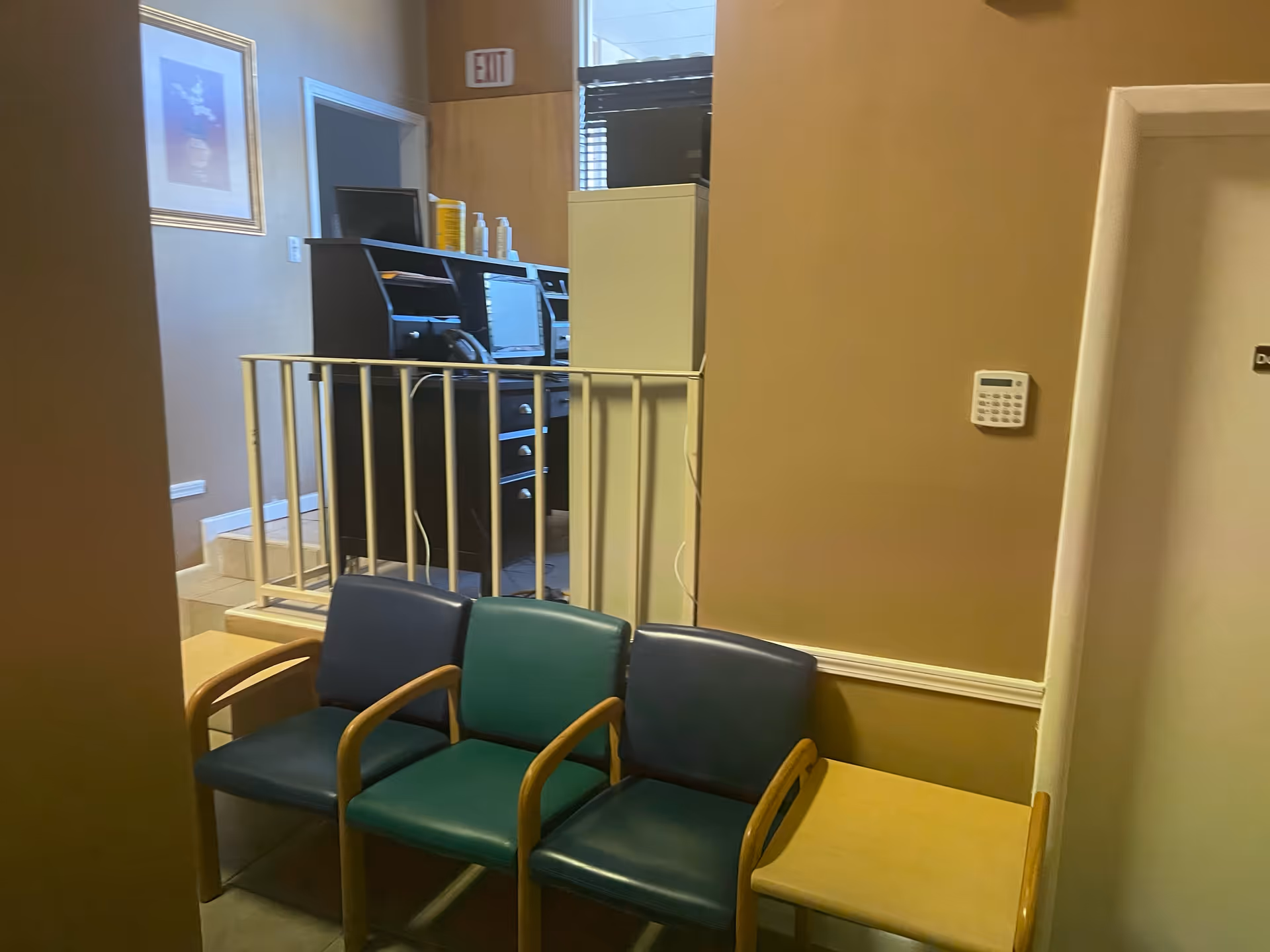 Indoor waiting area with teal chairs and a desk behind a railing in beige walls