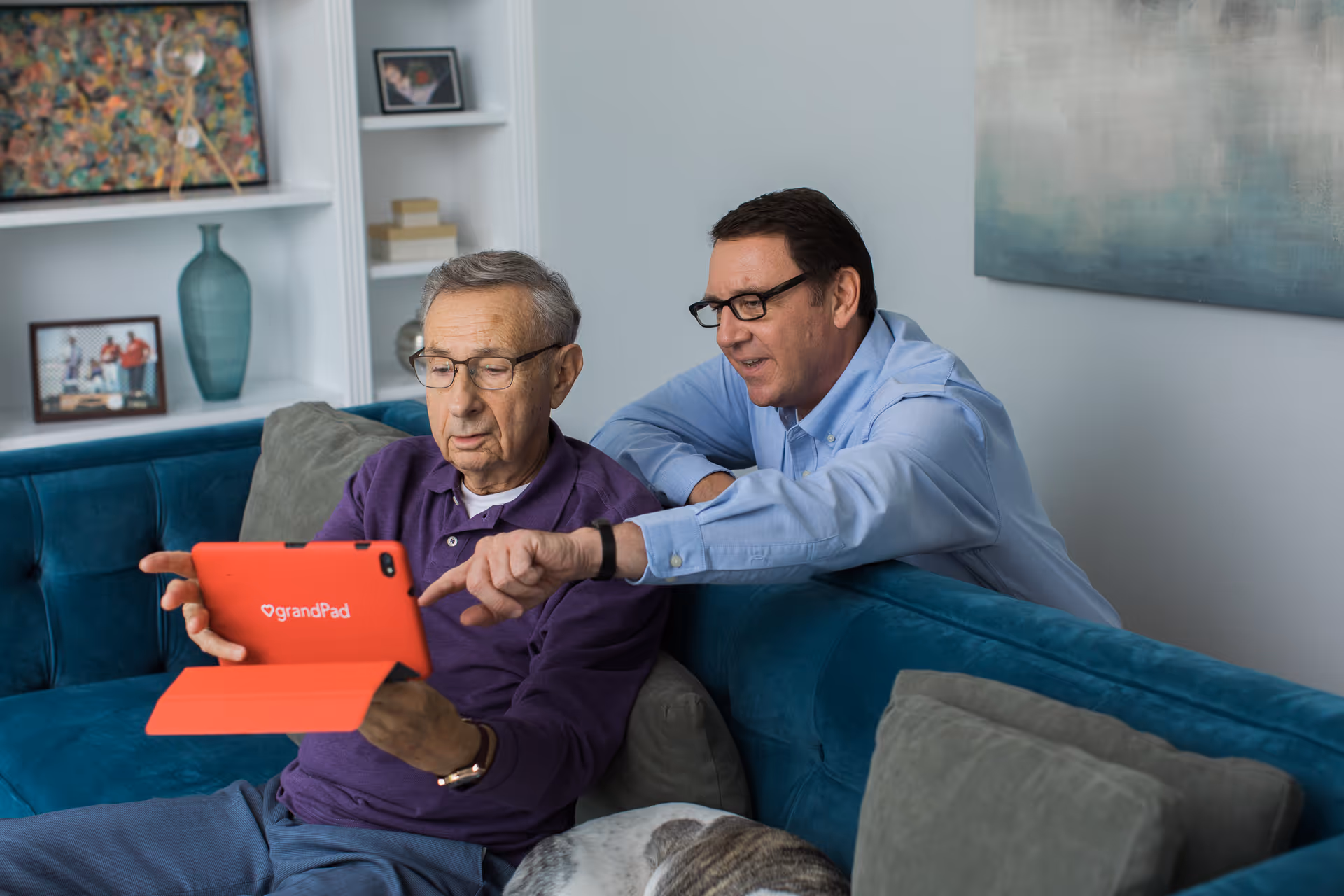 Elderly man and caregiver share a tablet on a teal living room sofa.